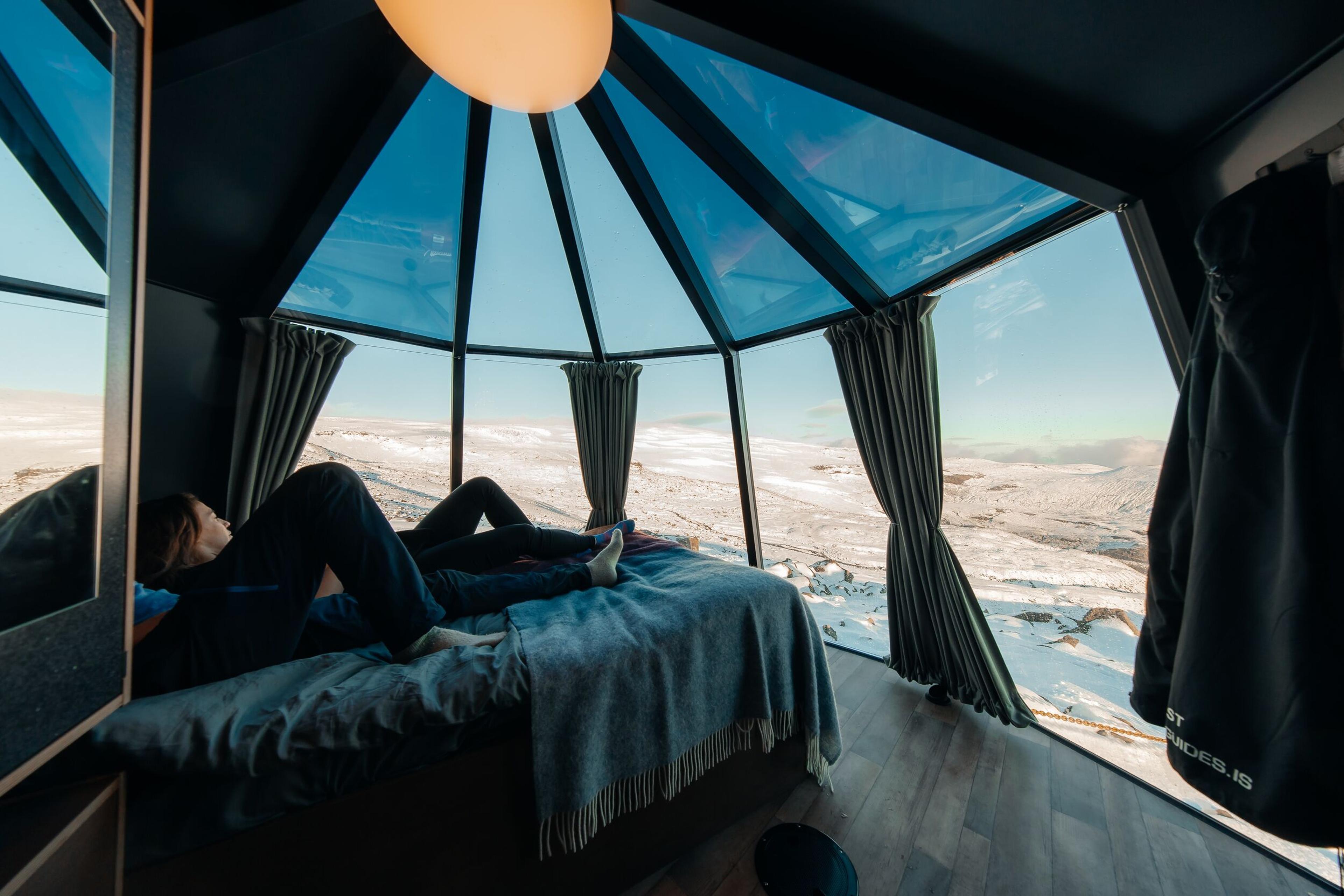 Two people relaxing on a bed in a glass-walled room overlooking a snowy landscape.