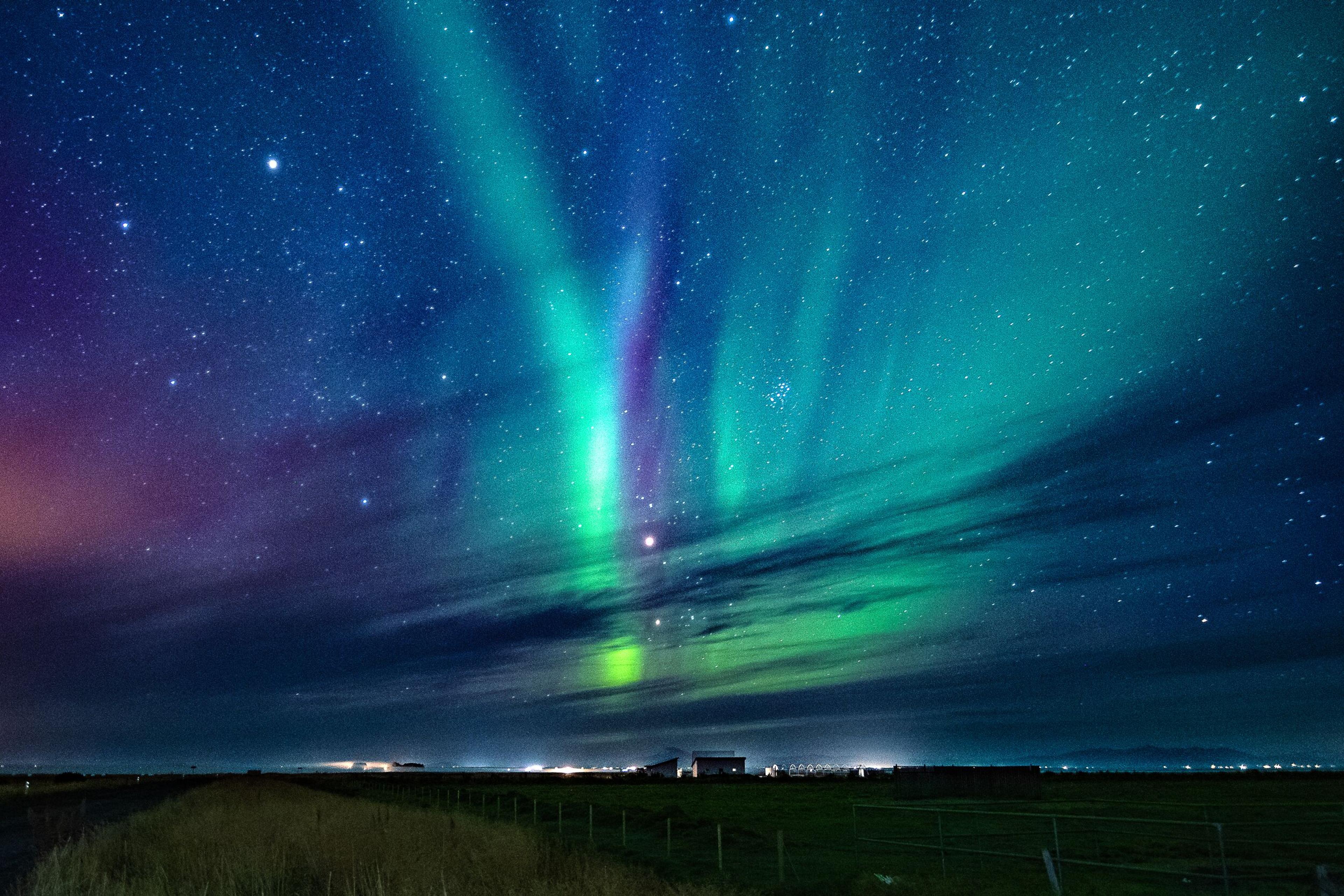 Northern Lights in green, blue, and purple illuminate a starry night sky over a dark landscape.
