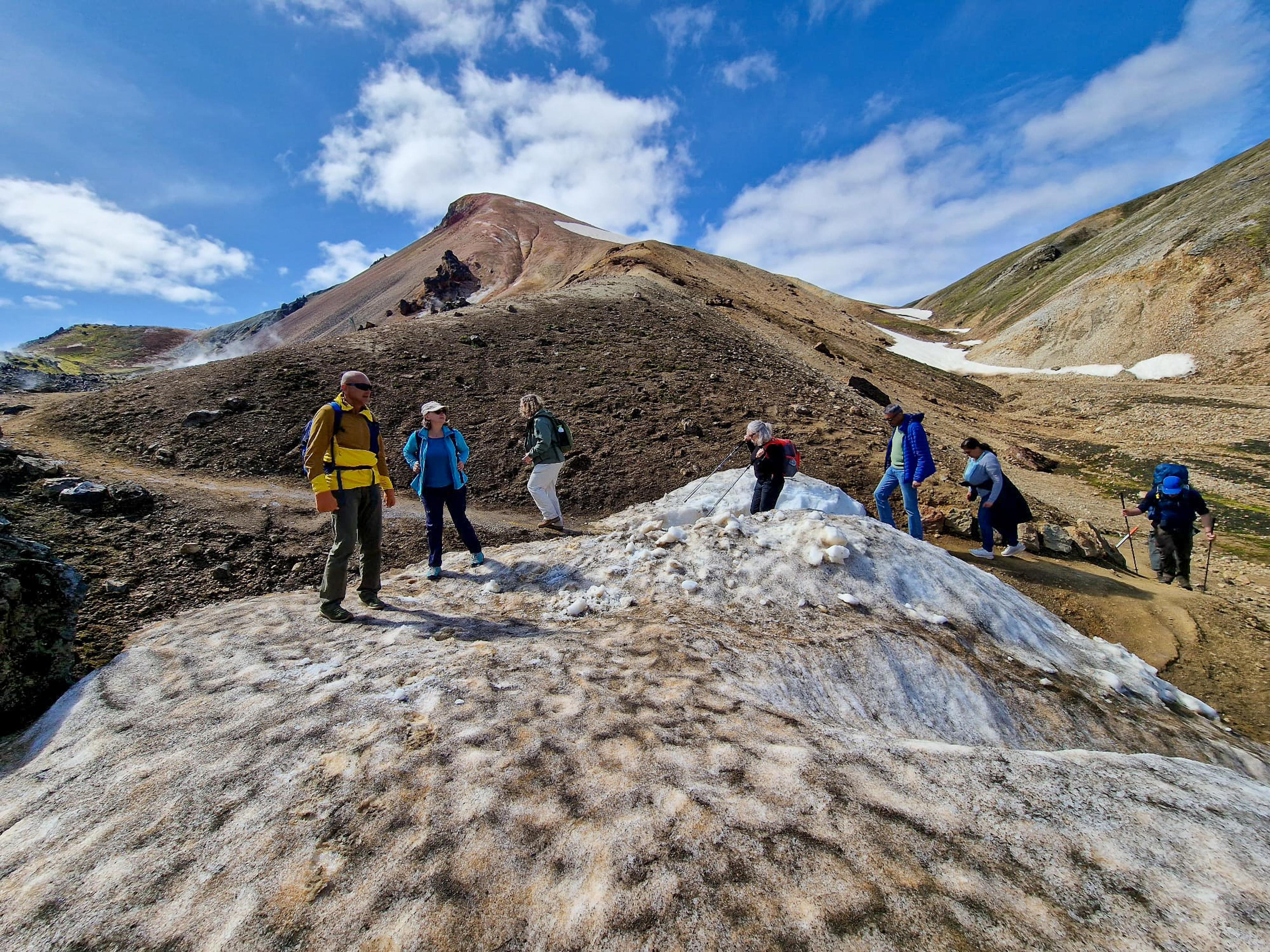 A group of hikers navigates a colorful volcanic landscape with patches of dirty snow under a blue sky.
