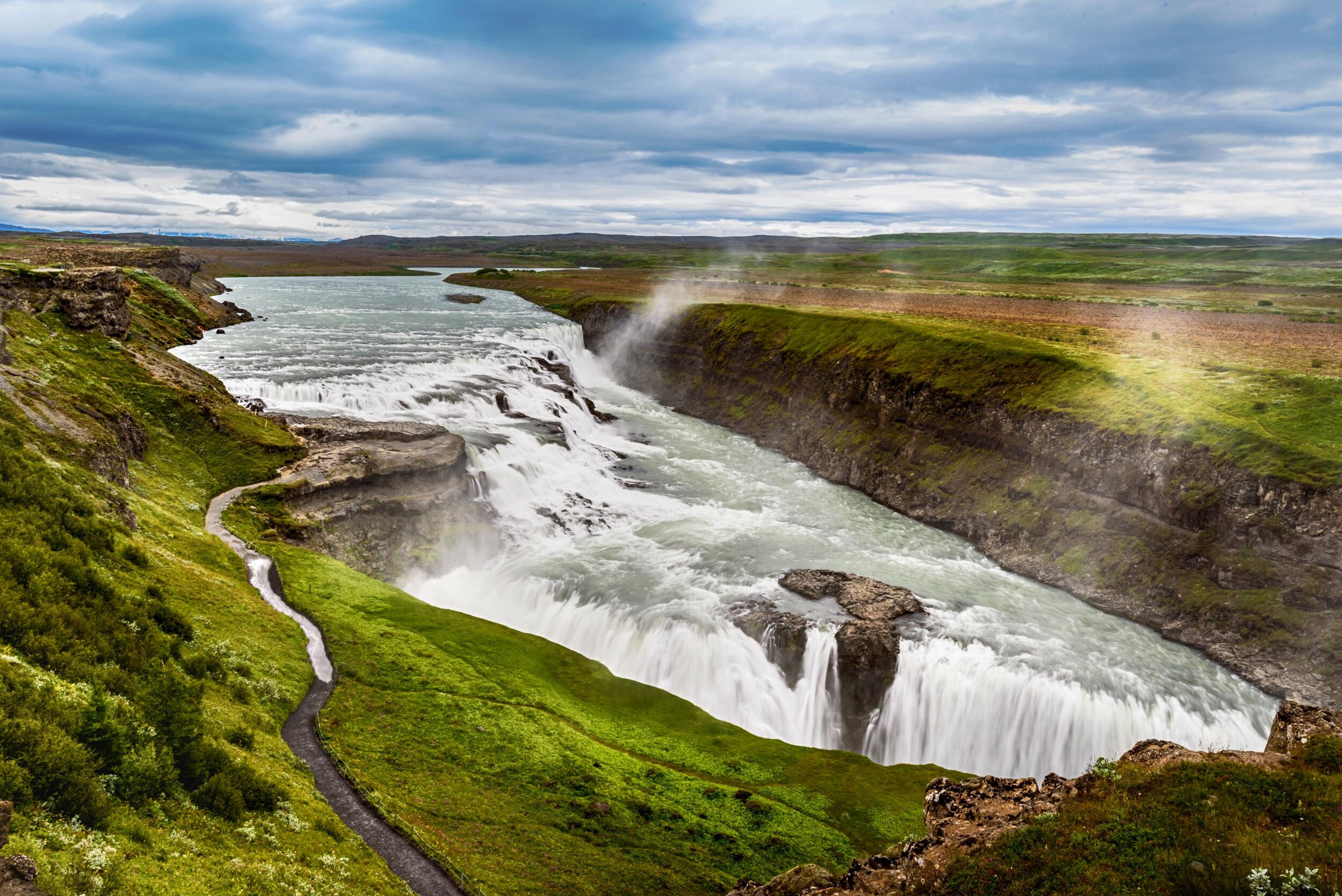 A wide, powerful waterfall cascades through a green gorge with a winding path on the left, under a cloudy sky.