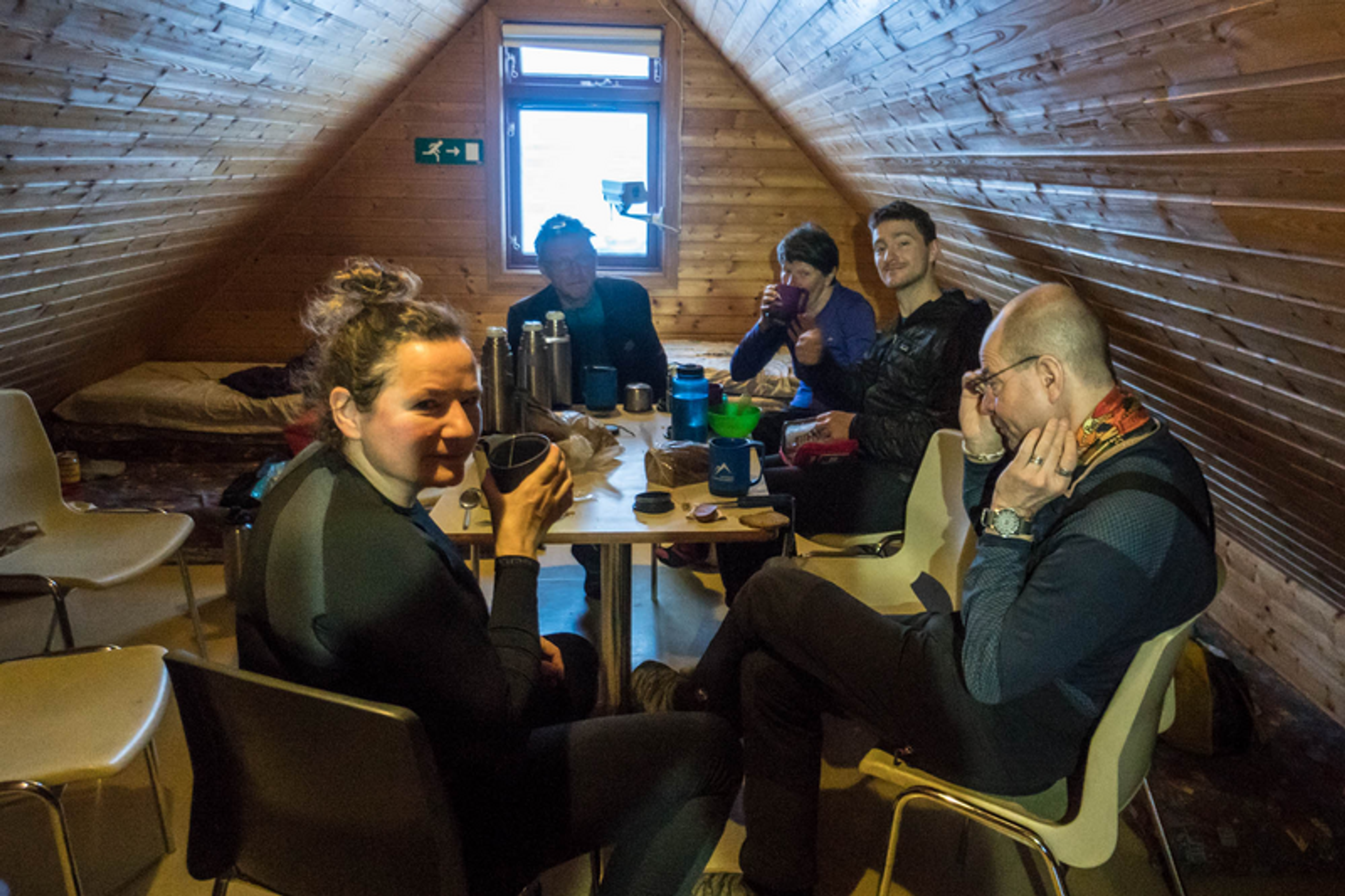 A group of people sitting around a wooden table drinking tea in a small wooden mountain hut