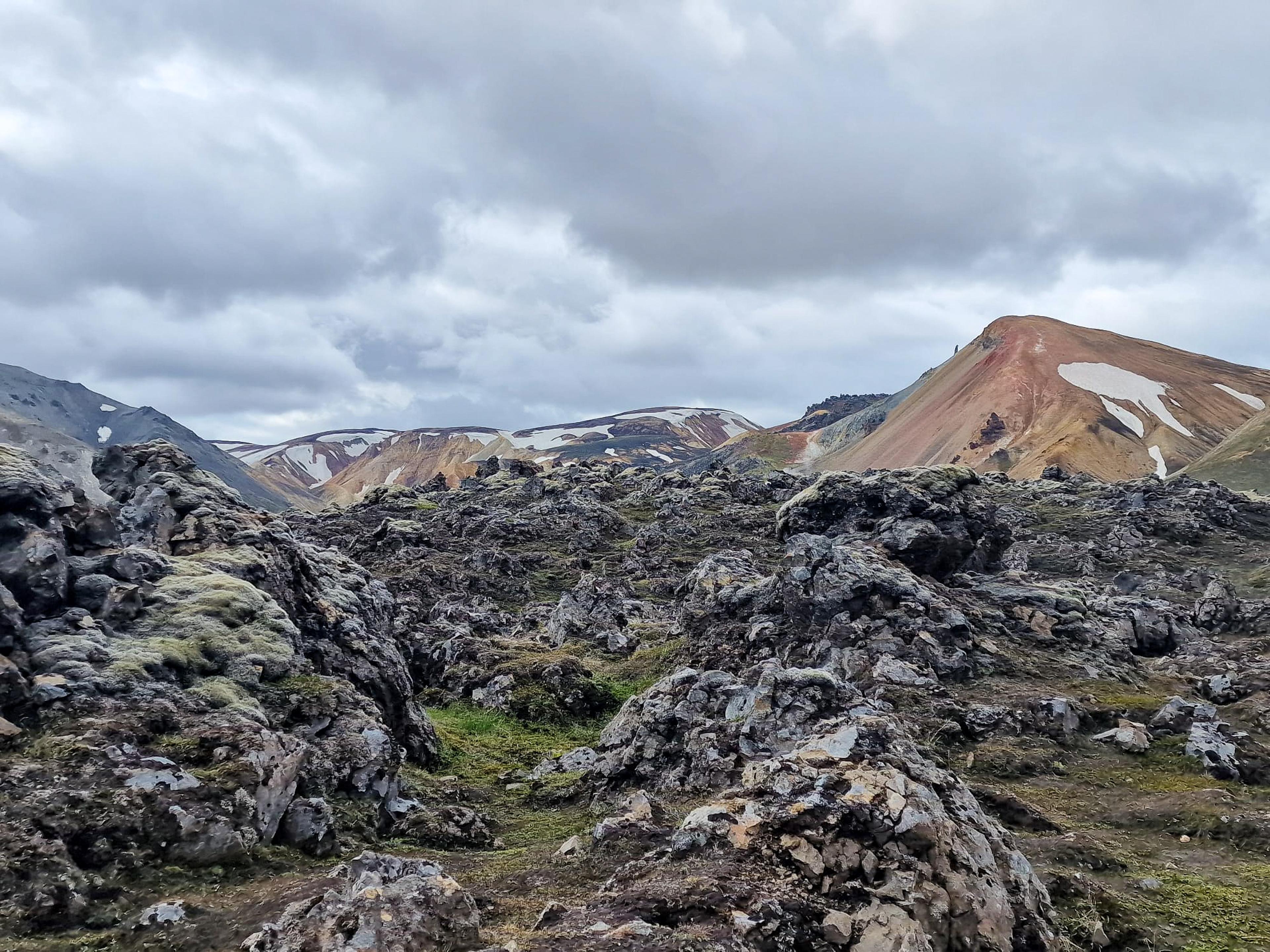 A rugged volcanic landscape with dark, moss-covered rocks and colorful mountains with snow patches under a cloudy sky.