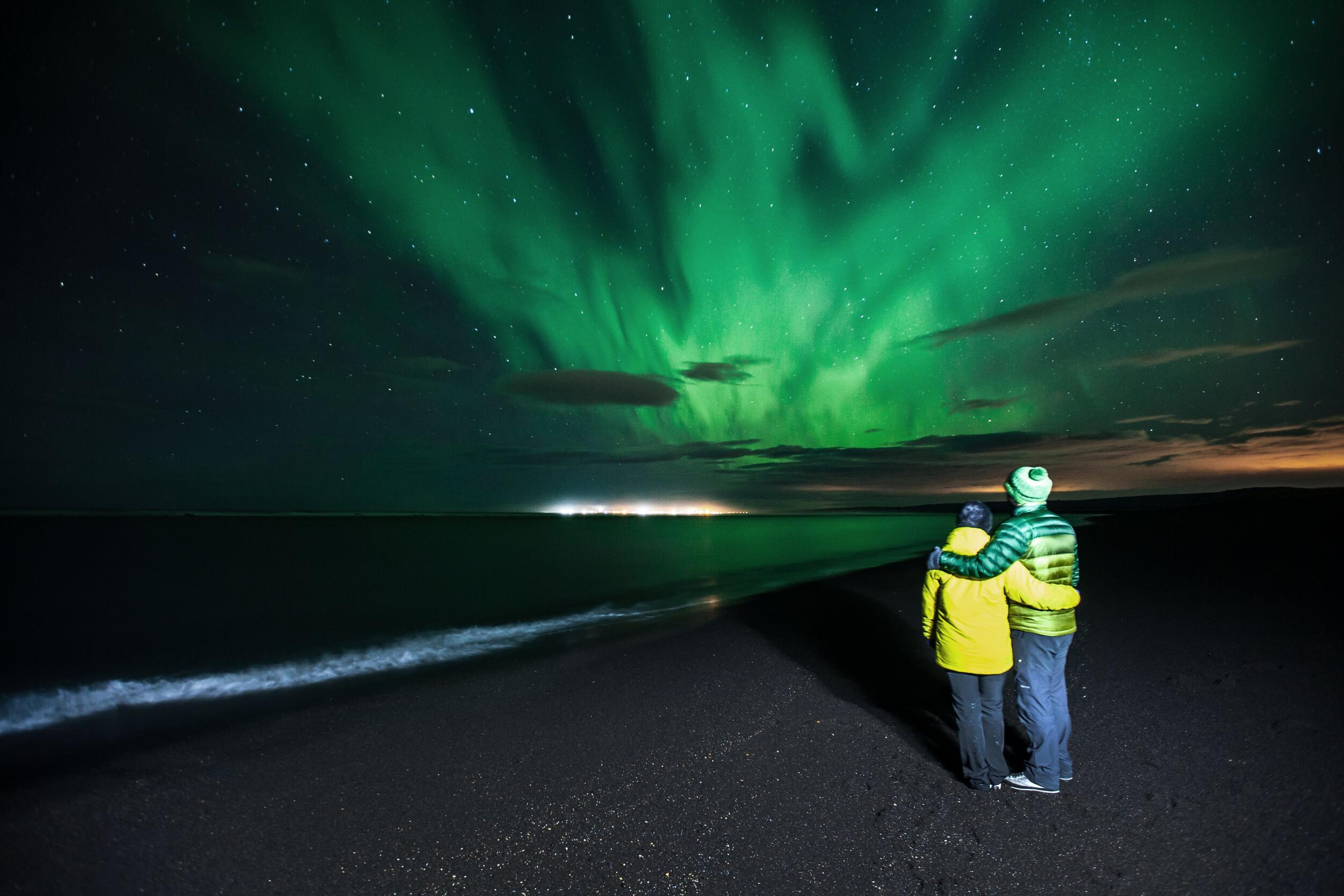 Two individuals, one wearing a green jacket and the other in yellow, embracing while watching the Northern Lights on a beach at night.