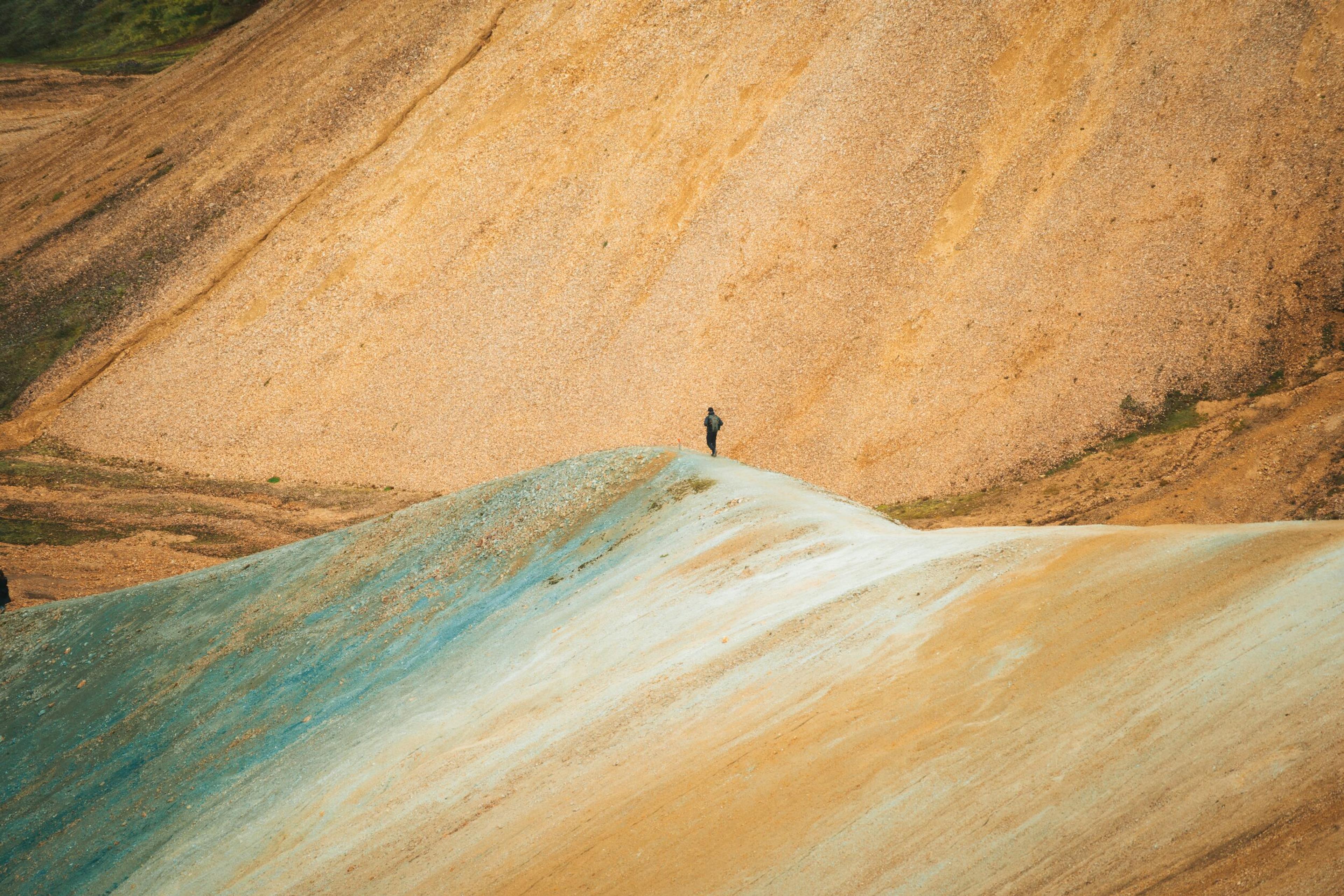 A lone person stands on a ridge of blue and sandy earth, against a backdrop of golden-brown mountains.