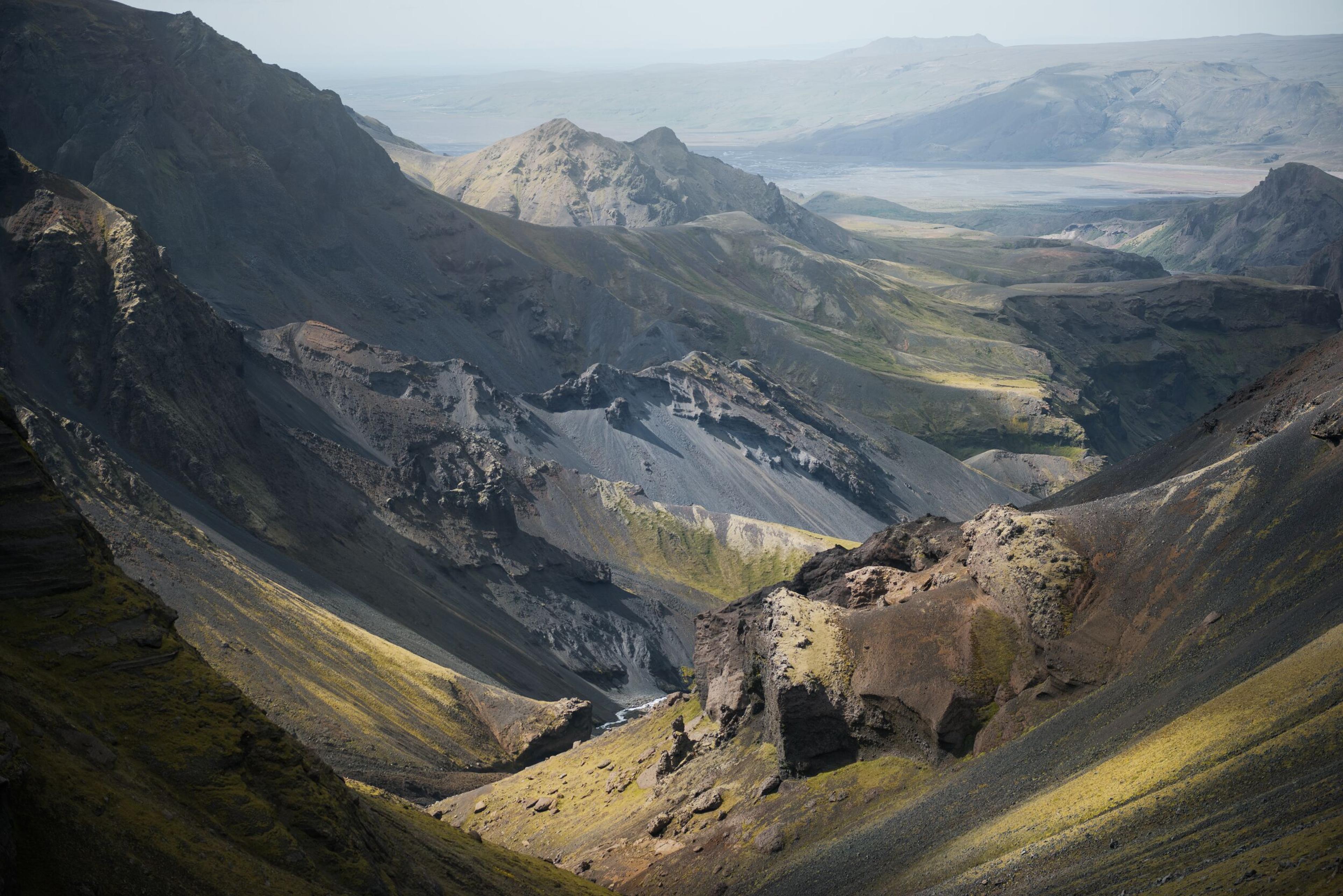 A deep, rugged volcanic valley with steep, dark gray slopes, patches of green vegetation, and a small river.