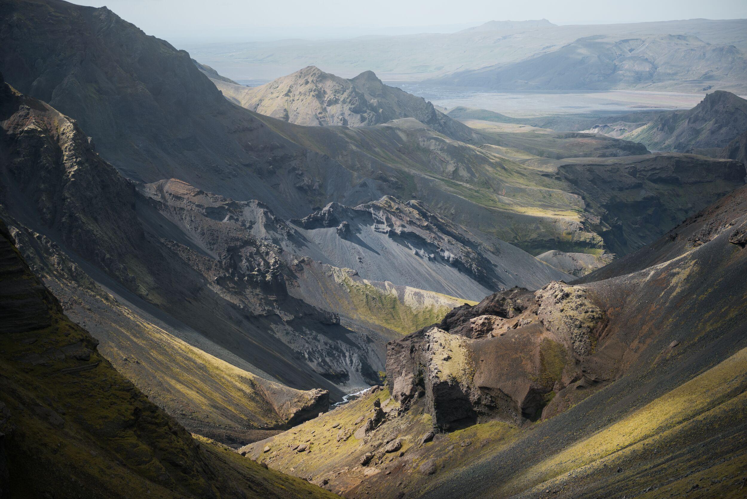 A deep, rugged volcanic valley with steep, dark gray slopes, patches of green vegetation, and a small river.