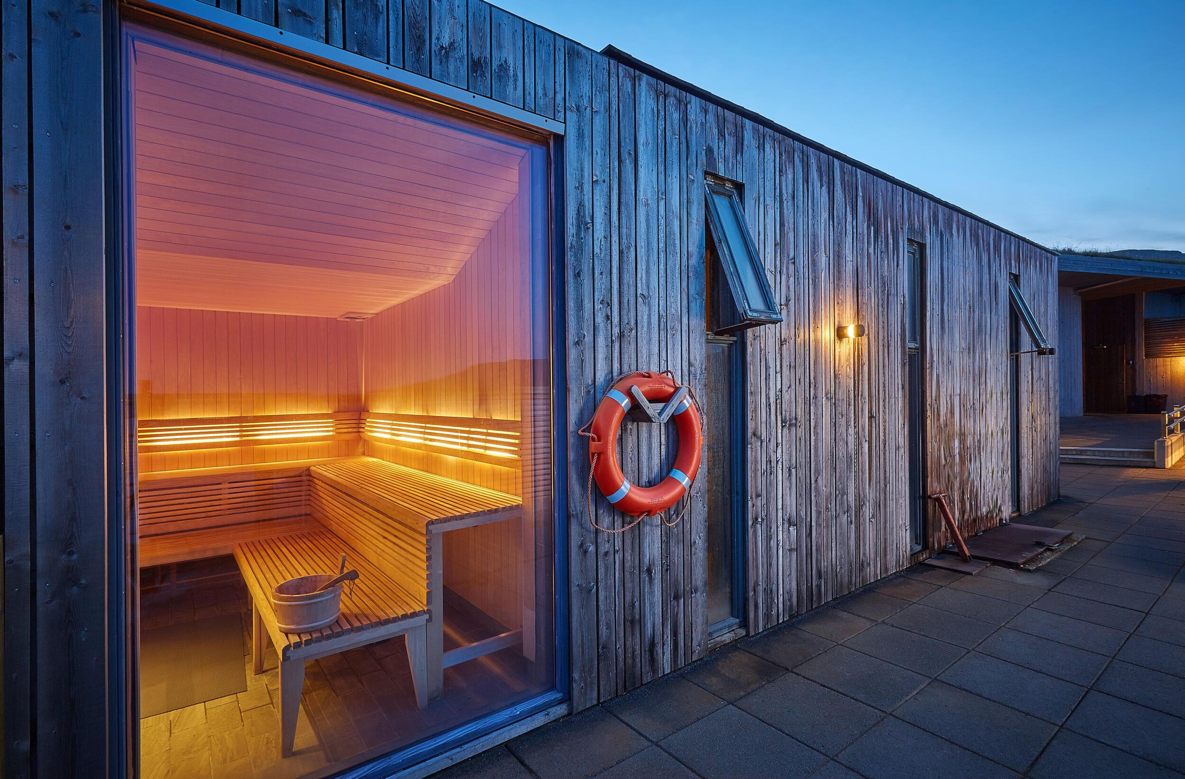 The warm glow of a sauna illuminates the evening as it invites relaxation within its wooden walls, complemented by a rustic exterior and a lifebuoy hanging, suggesting a location near water.