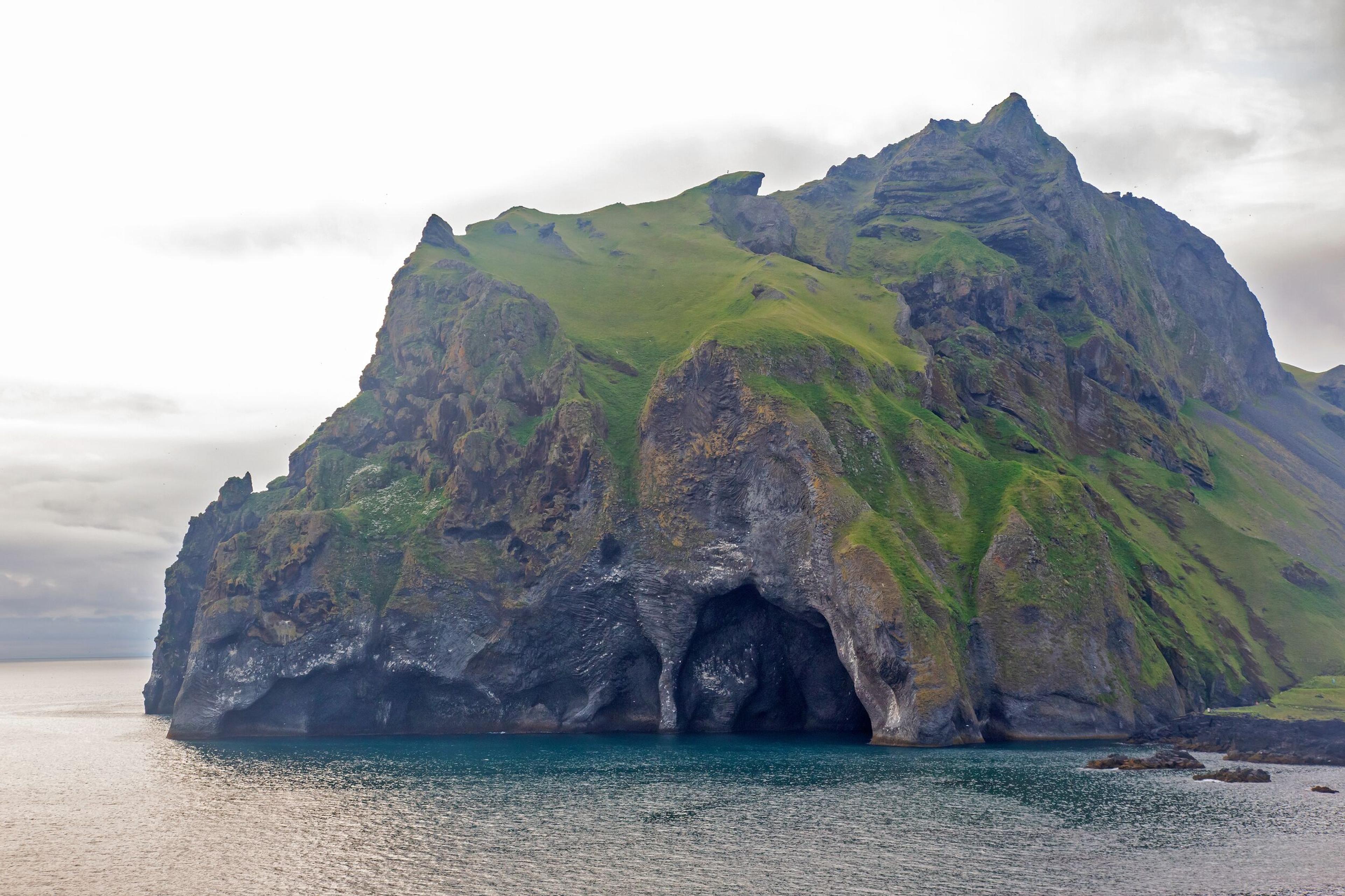 A rocky island with a green top and large sea caves, in blue water under a grey sky.