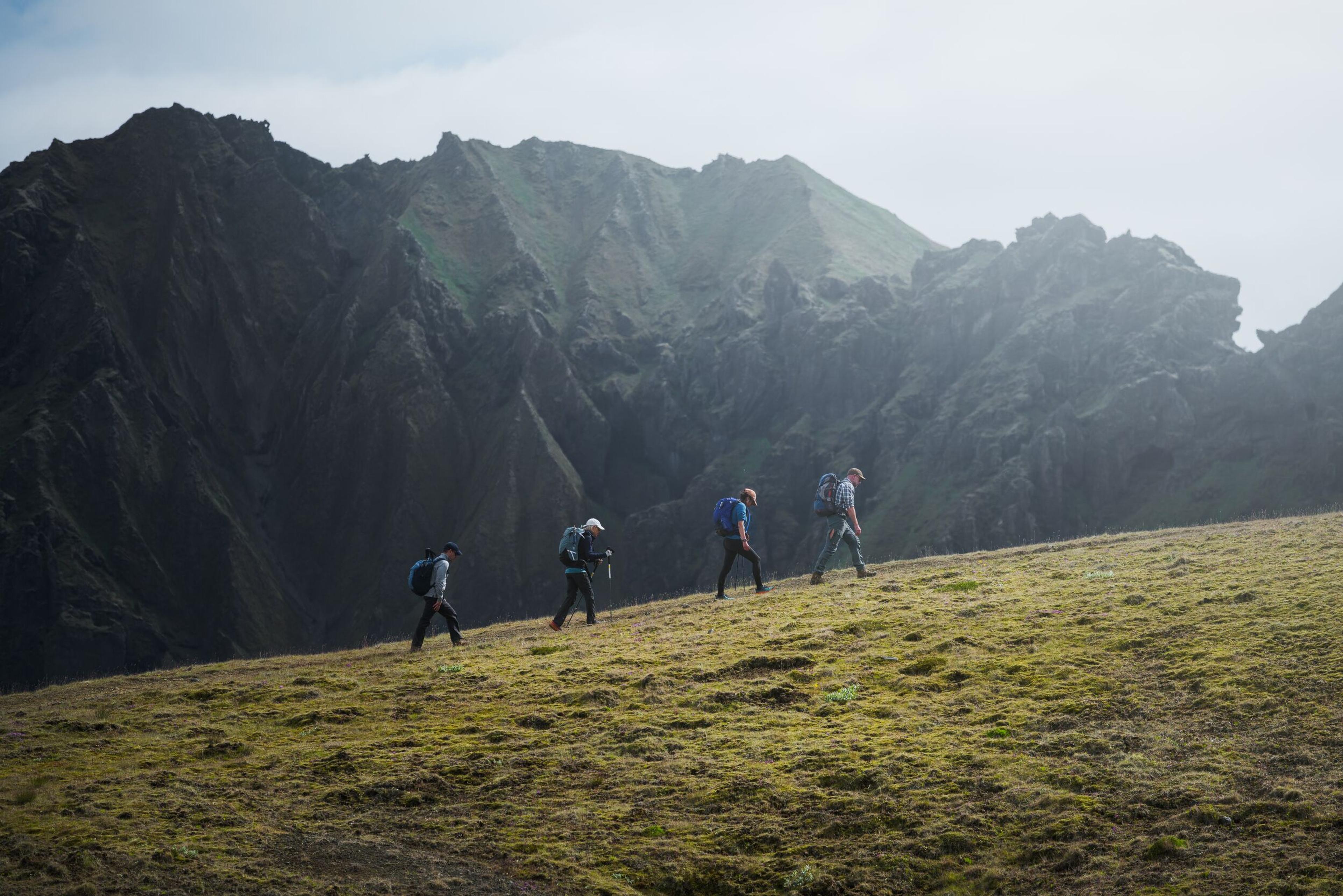 Four hikers walking across grassy slopes with steep, rocky cliffs towering beside them in Thorsmörk.