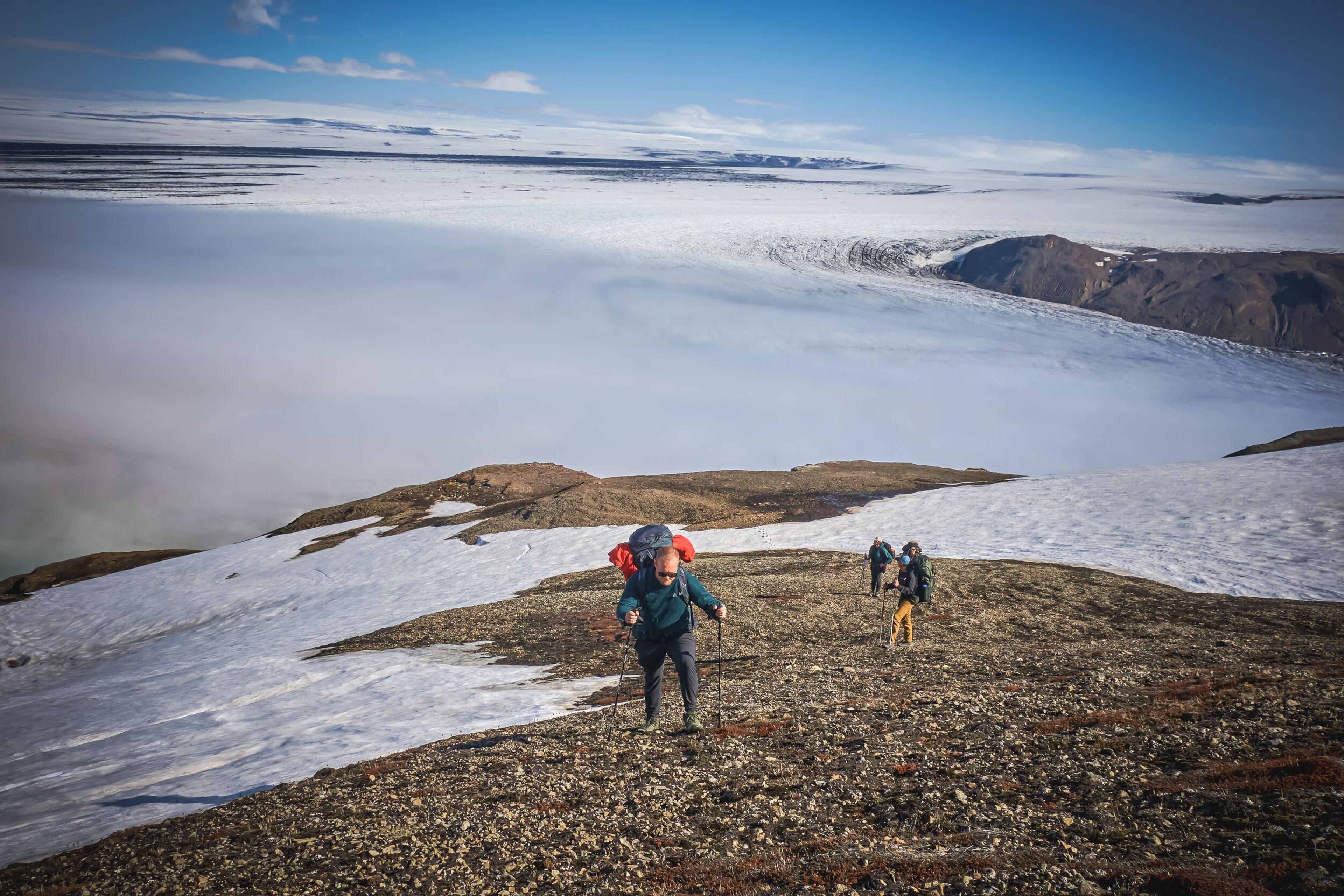 Three hikers ascend a rocky, snow-dusted slope above a cloud-filled valley and distant glaciers.