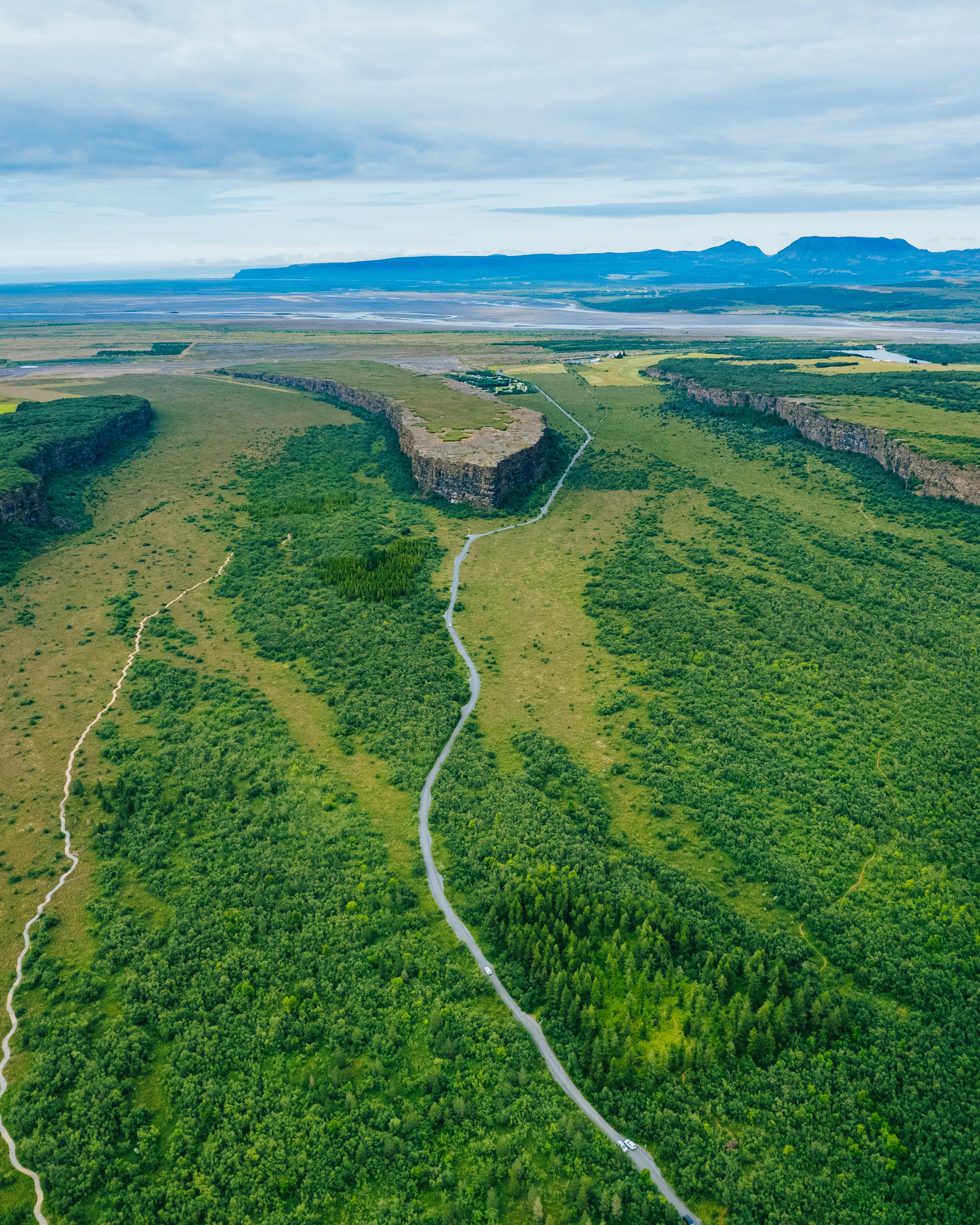 Aerial view of Ásbyrgi Canyon, showing a meandering trail through lush greenery, with a backdrop of distant flat-topped mountains under a cloudy sky.