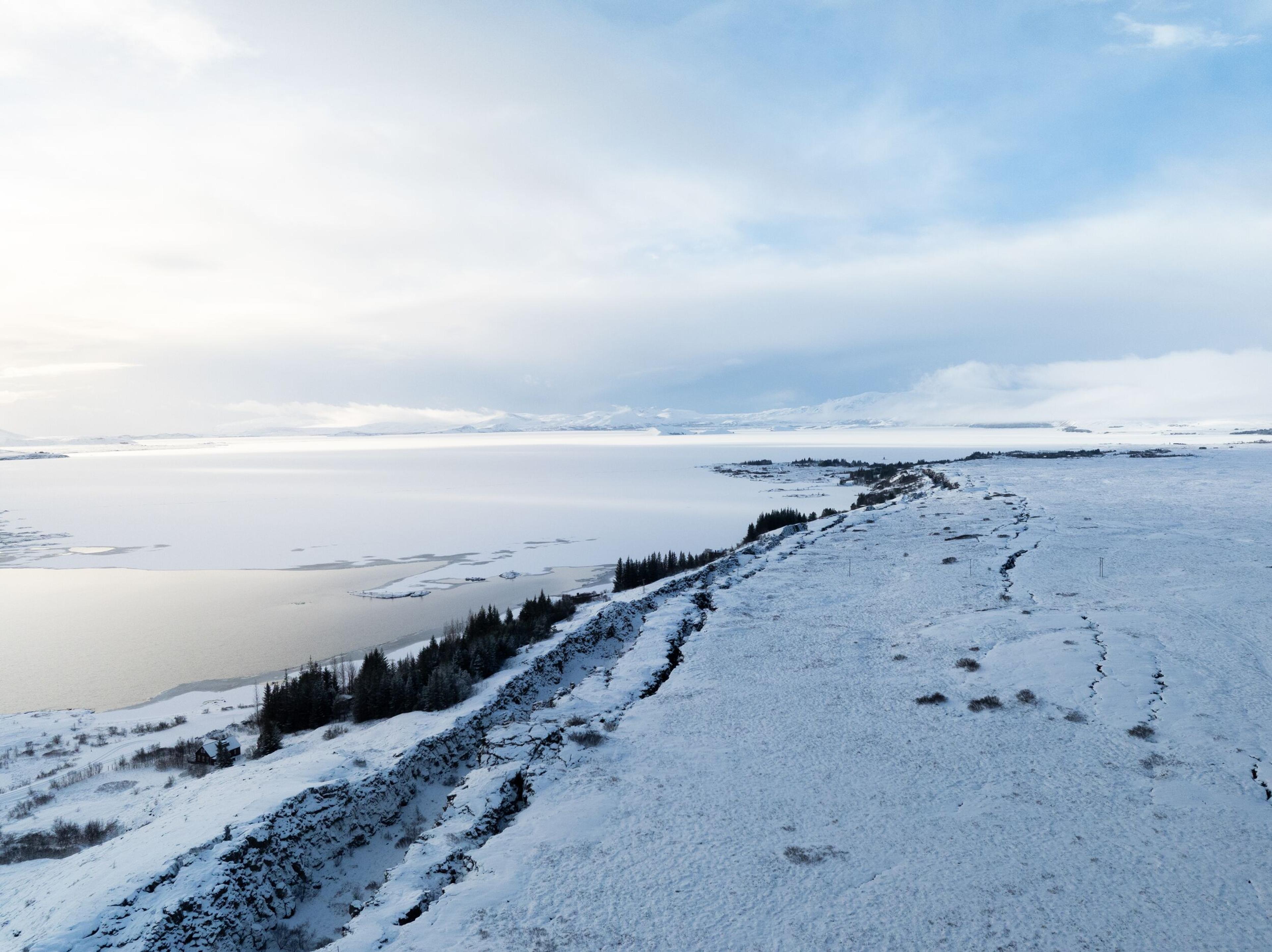 A vast winter landscape with a snow-covered fissure, a partially frozen lake, and dark evergreen trees.