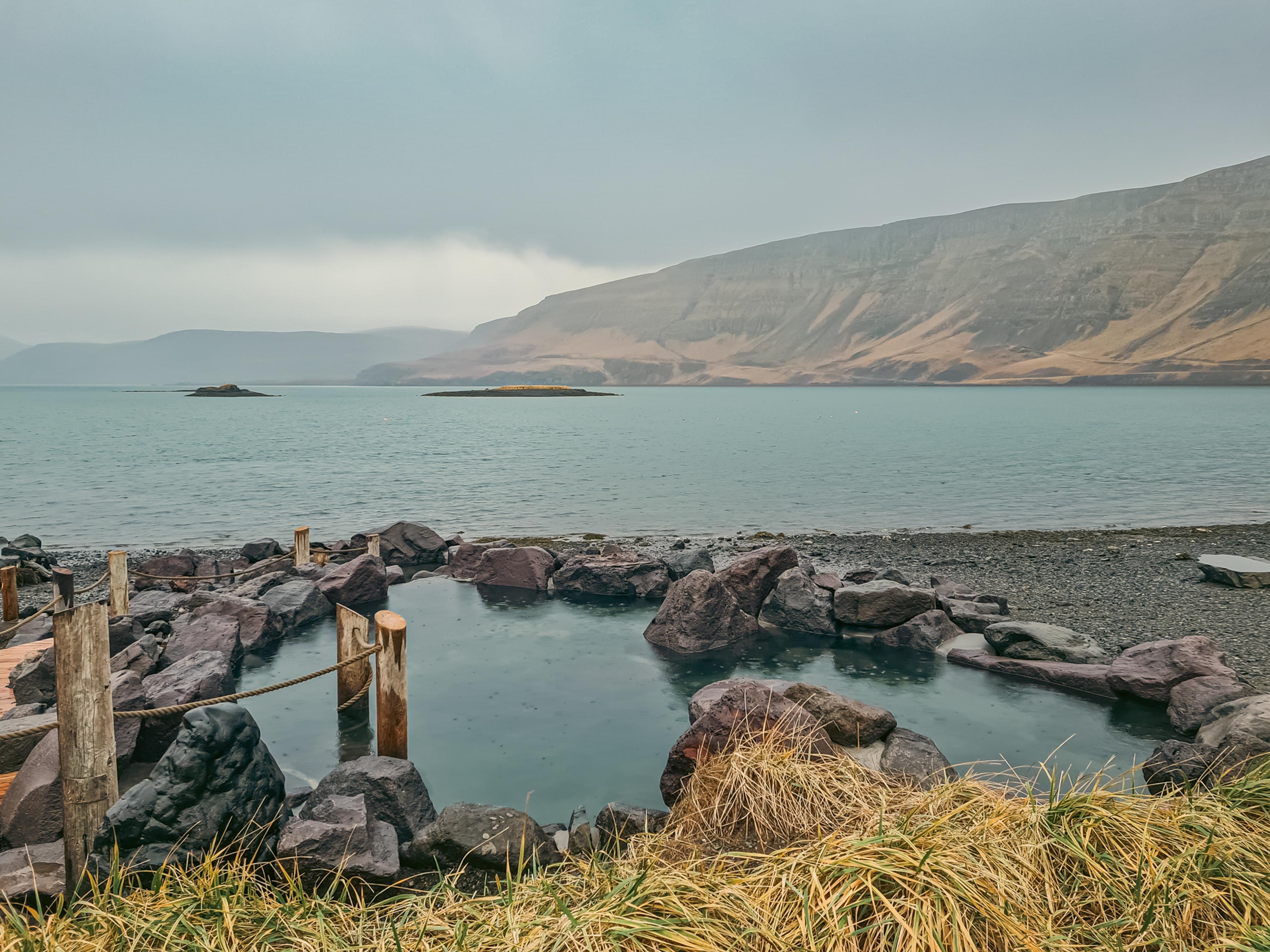 A natural hot spring pool on a rocky shore, with a calm body of water and mountains in the background under a cloudy sky.