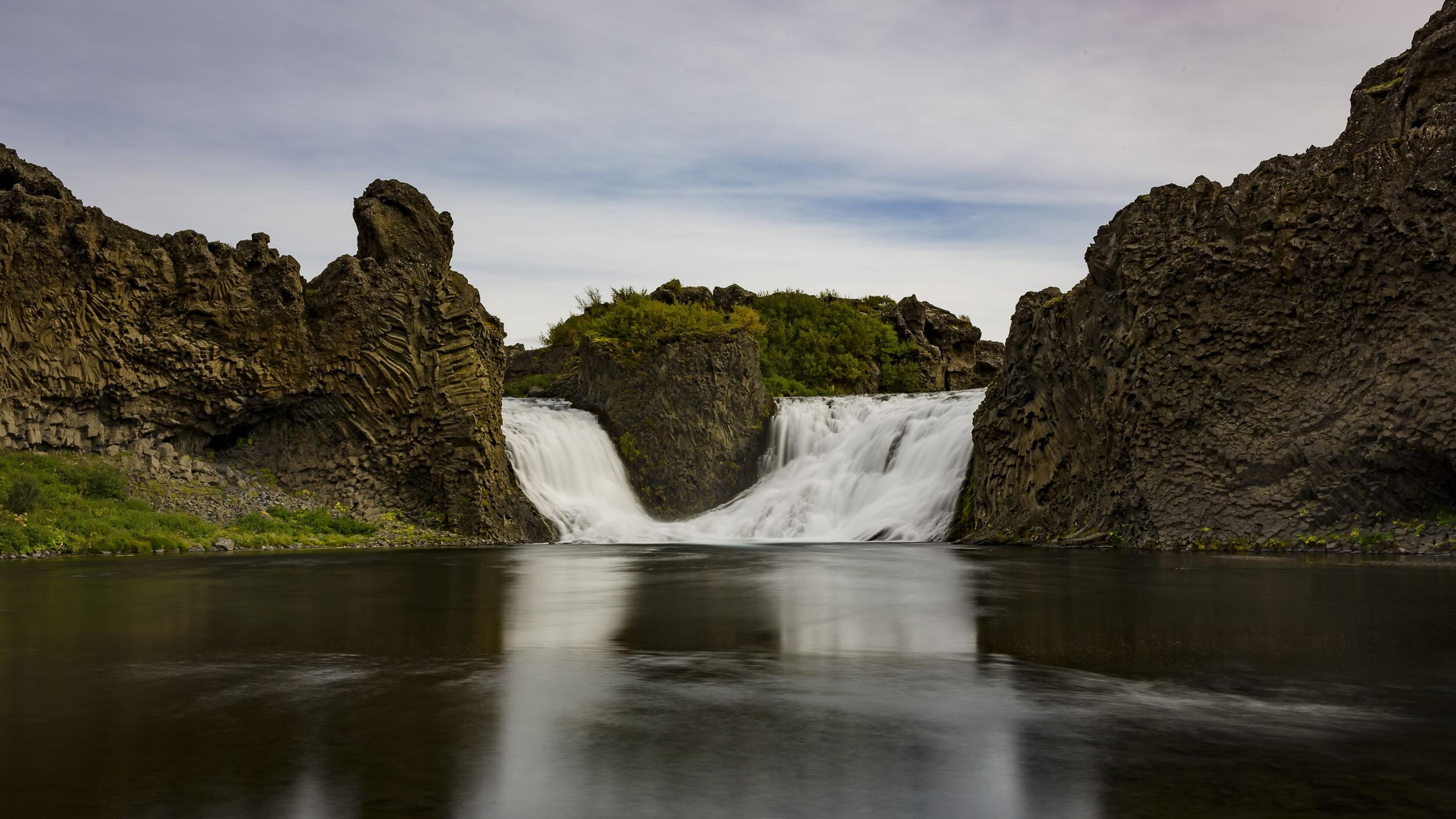 A wide waterfall with smooth, white water flows between dark, textured rock formations into a calm, reflective river.