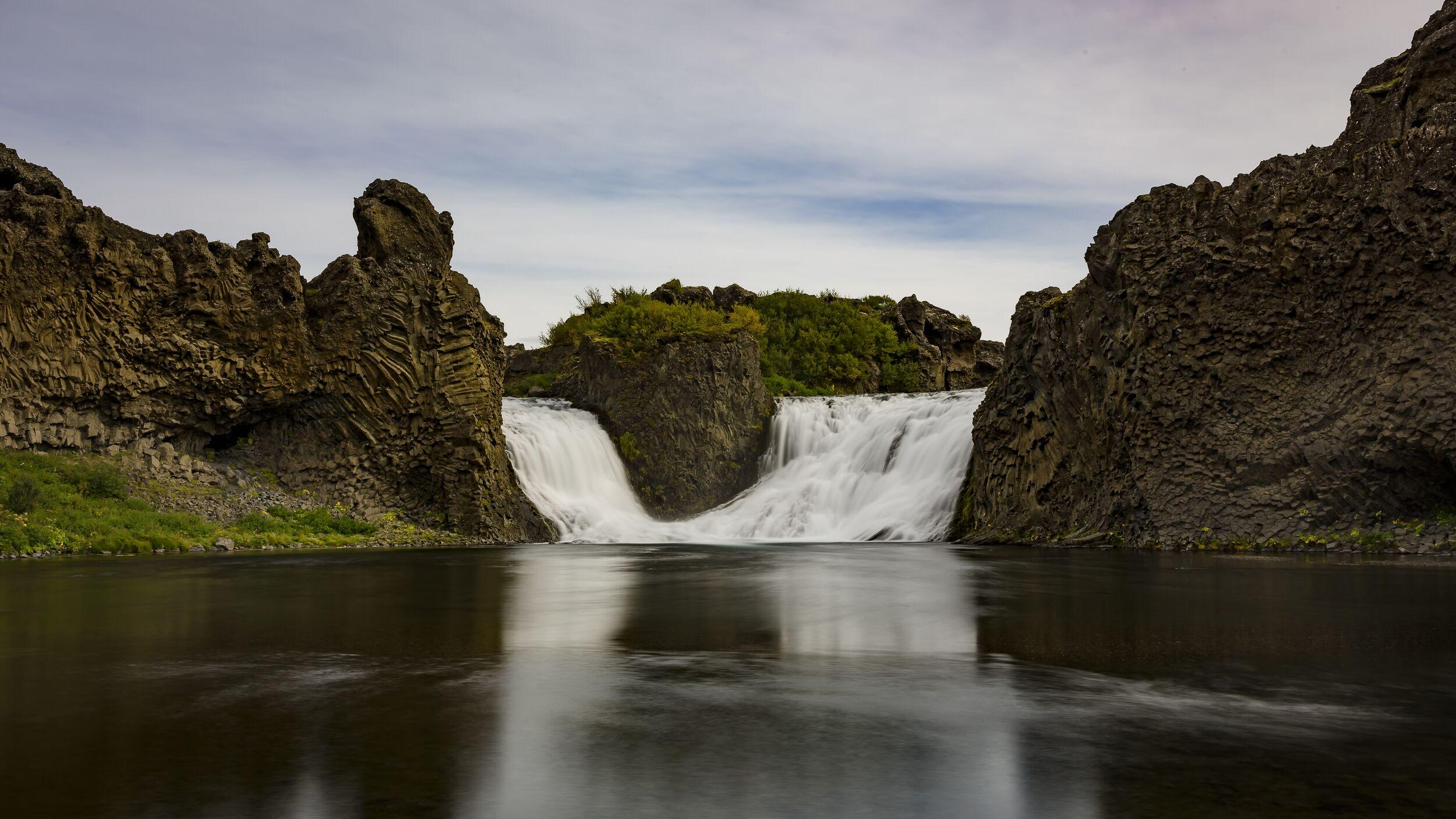 A wide waterfall with smooth, white water flows between dark, textured rock formations into a calm, reflective river.