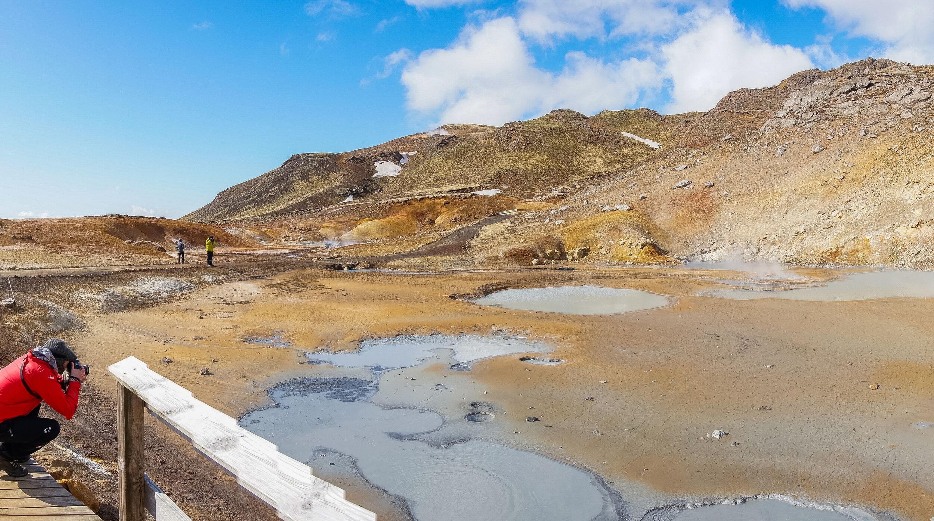 Seltún geothermal area, featuring bubbling mud pools, steaming vents, and colorful mineral deposits, with a wooden boardwalk 