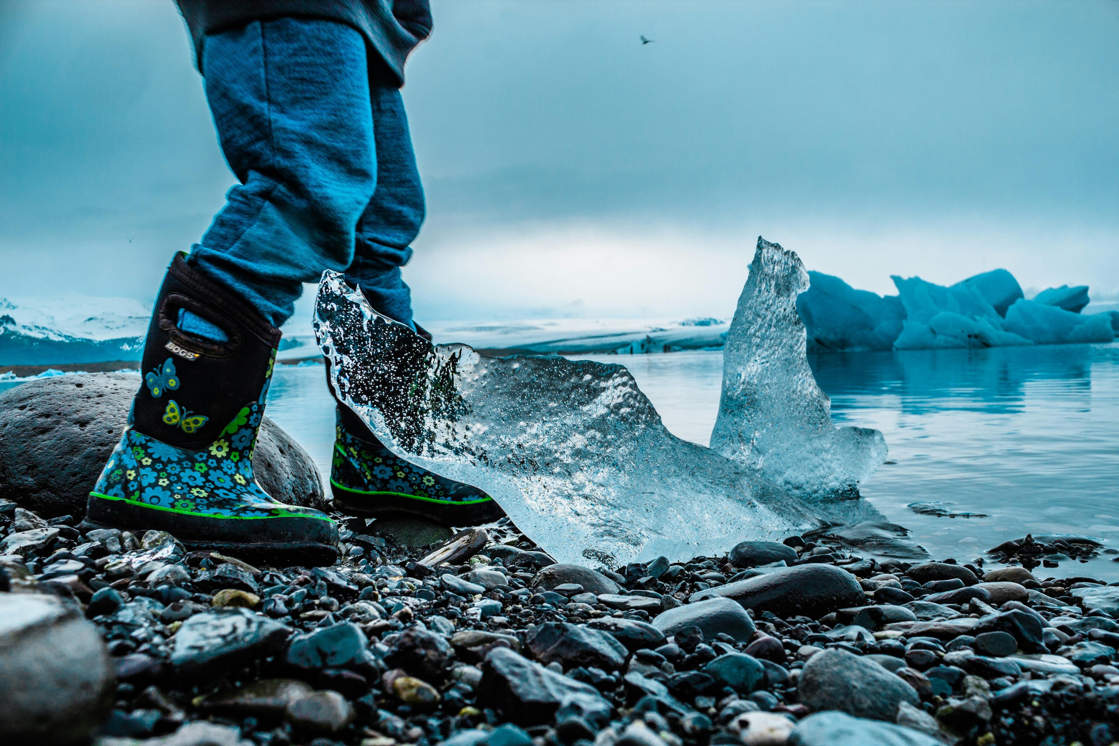 A child wearing colorful rain boots standing next to a clear ice chunk on a rocky shore in Iceland, with glaciers in the background.