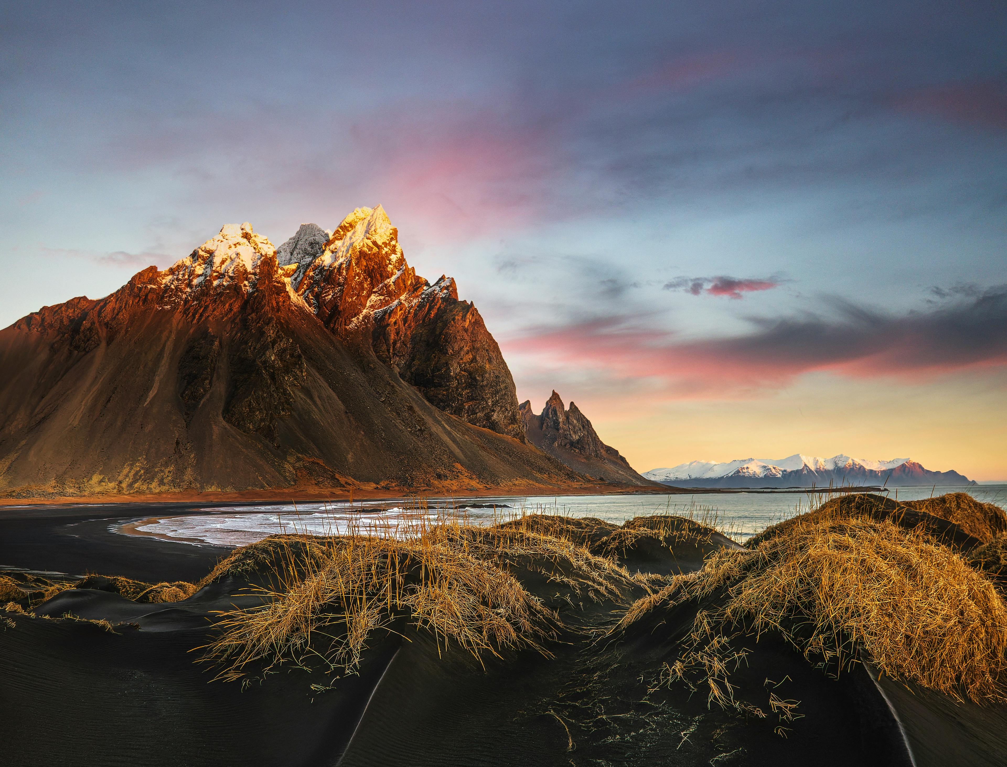This image showcases the breathtaking Stokksnes Peninsula in Iceland, with golden grasses covering black sand dunes in the foreground. The majestic Vestrahorn mountain, illuminated by warm sunset hues, rises dramatically against the vivid pink and blue sky