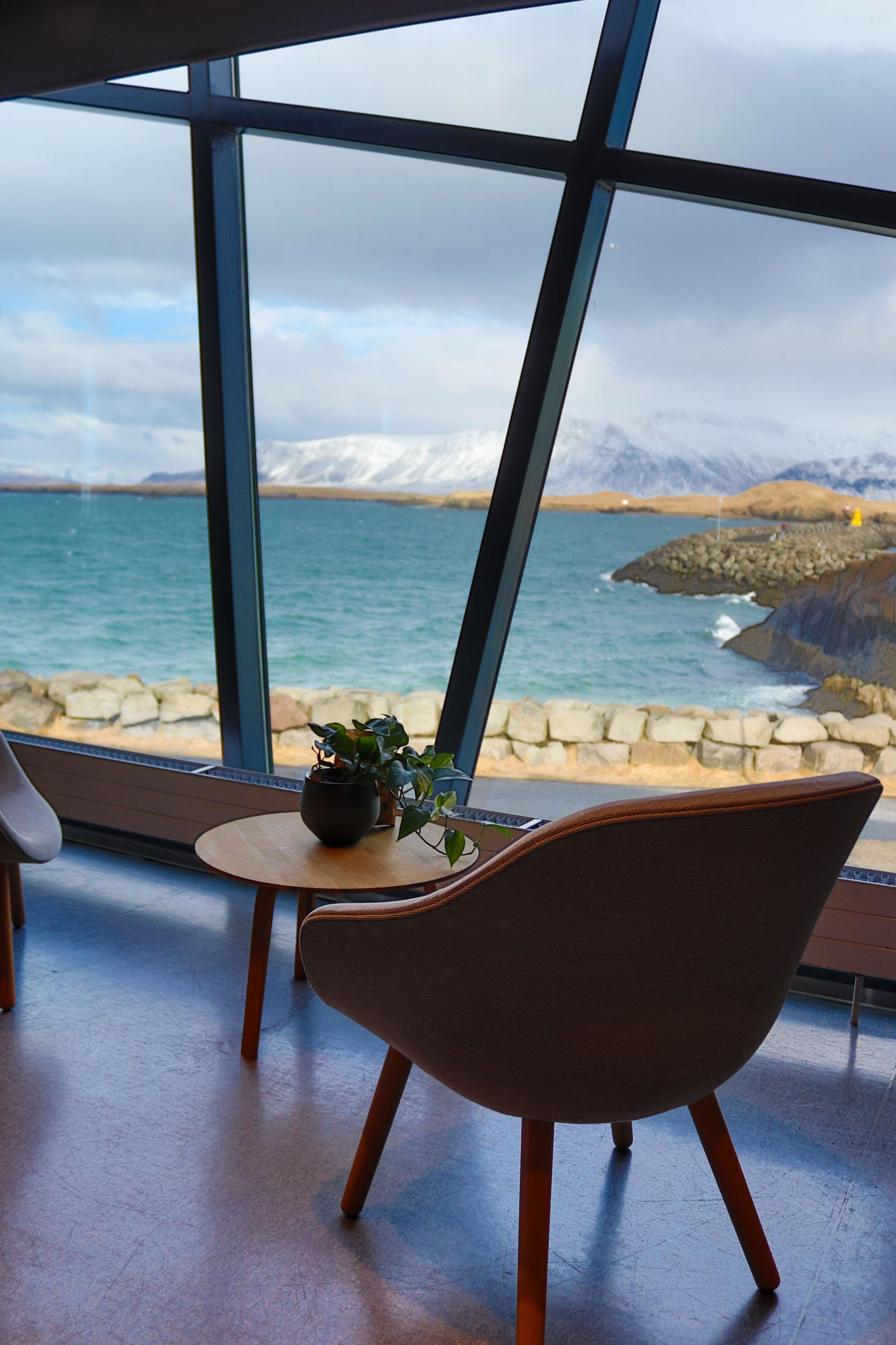A room with modern furnishings including two chairs and a table, view through the window with a focus on a table set with two chairs, a small plant in the center, and a pair of glasses, suggesting a cozy spot for a meeting or relaxation with the same picturesque view of the sea and mountains.
