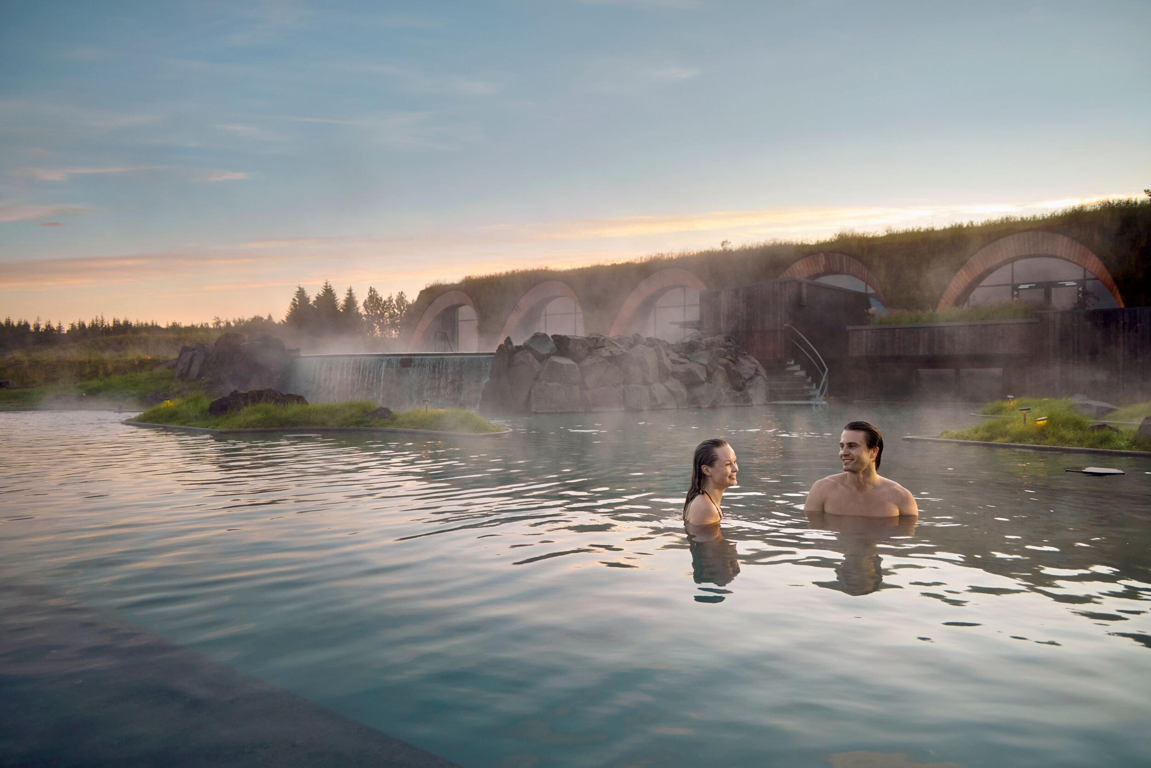 A couple smiles in a steaming geothermal pool with grass-roofed buildings and a waterfall in the background.