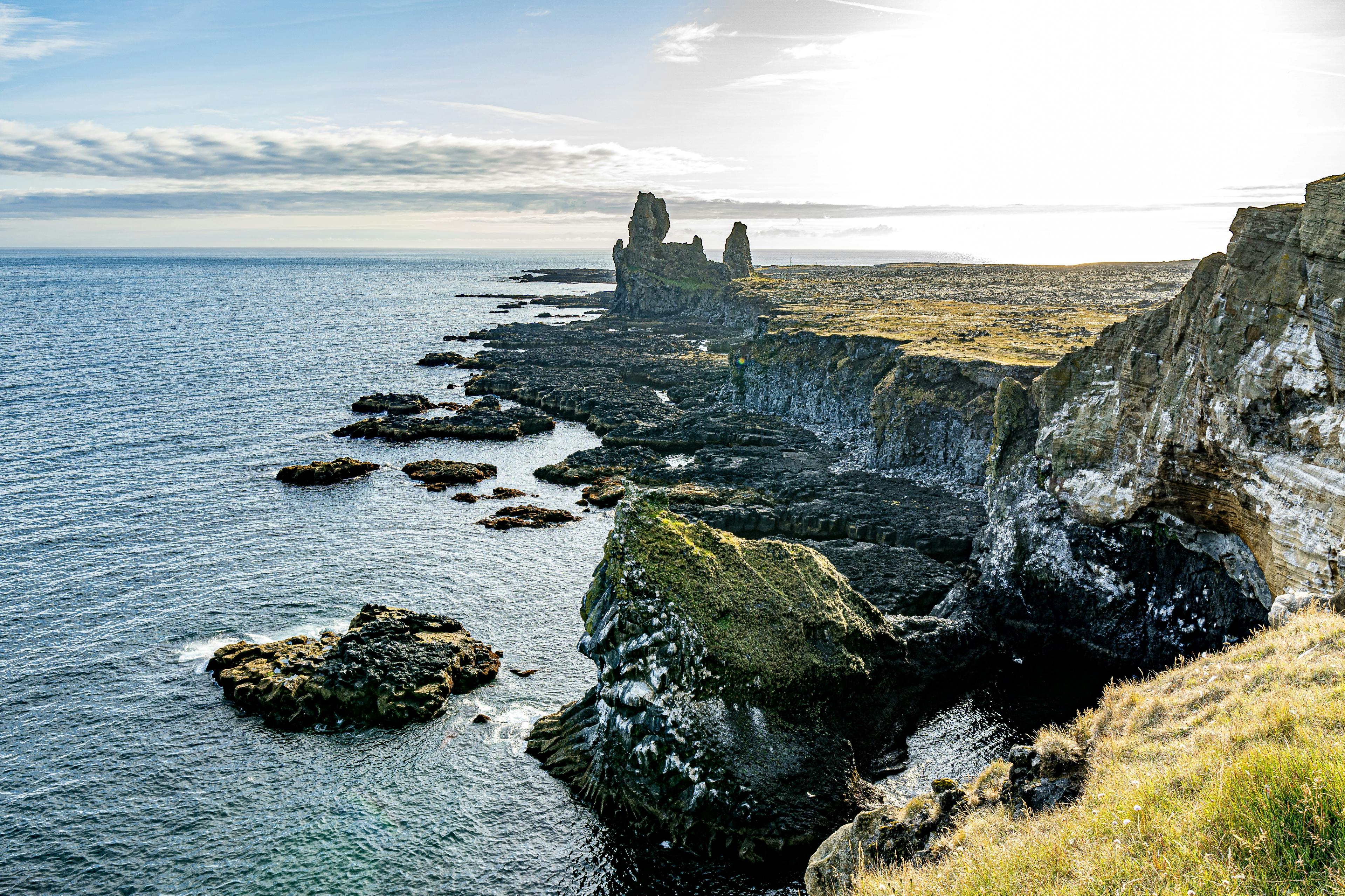 The rugged Longrangar sea cliffs in Iceland stretch along the coastline, with dramatic rock formations jutting into the ocean. The black volcanic rock contrasts with patches of green moss and grass, while the calm sea reflects the soft glow of the sky. The striking twin sea stacks in the distance stand as remnants of ancient lava flows, adding to the dramatic landscape.