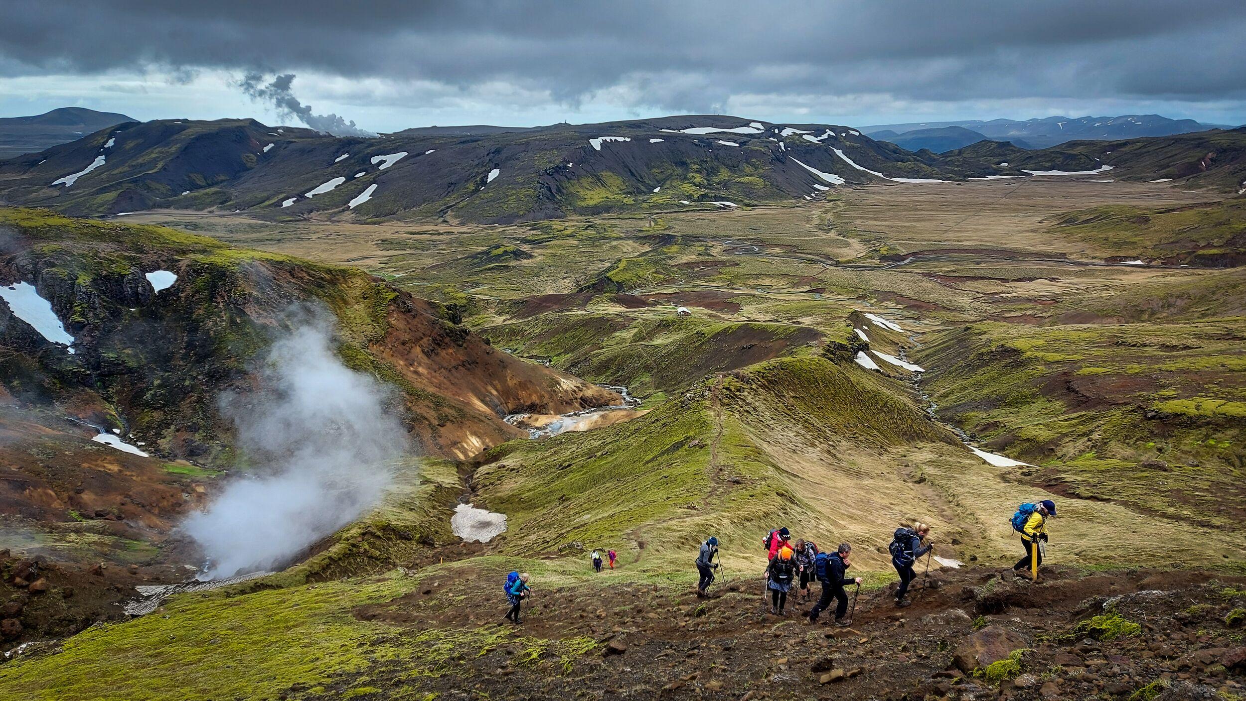 A group of hikers ascends a mossy hill overlooking a steaming geothermal valley and snow-dusted mountains.