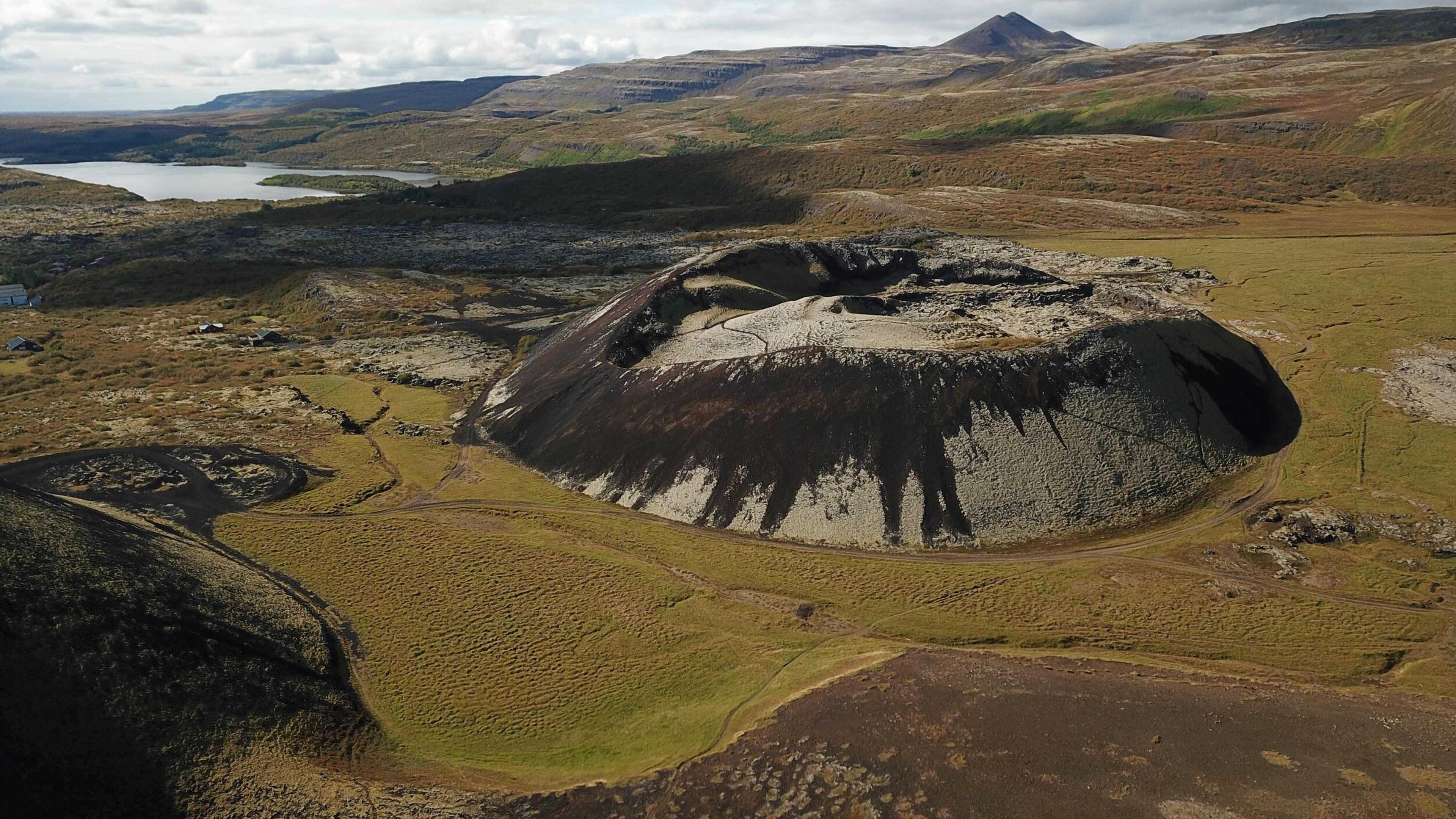 Aerial view of Grábrók crater, a volcanic cone surrounded by vast Icelandic landscapes with patches of green and brown, and distant lakes and mountains.