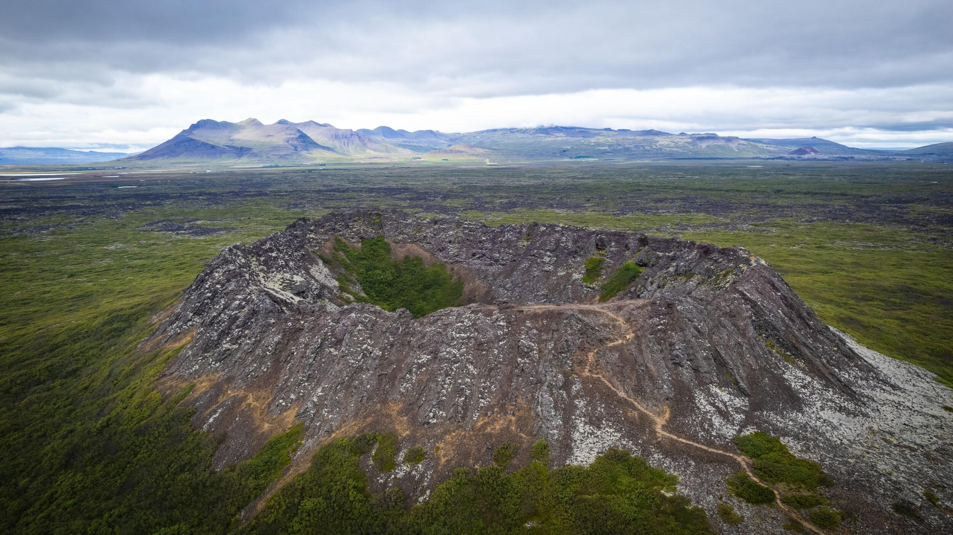 Aerial view of a volcanic crater with a path leading to its rim, surrounded by green and dark volcanic landscape under a cloudy sky.