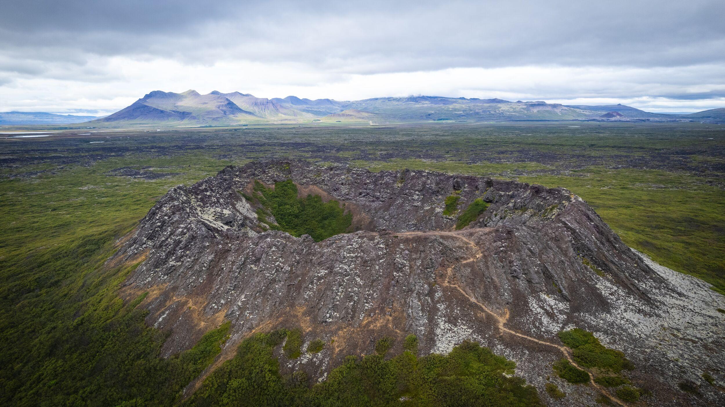Aerial view of a volcanic crater with a path leading to its rim, surrounded by green and dark volcanic landscape under a cloudy sky.