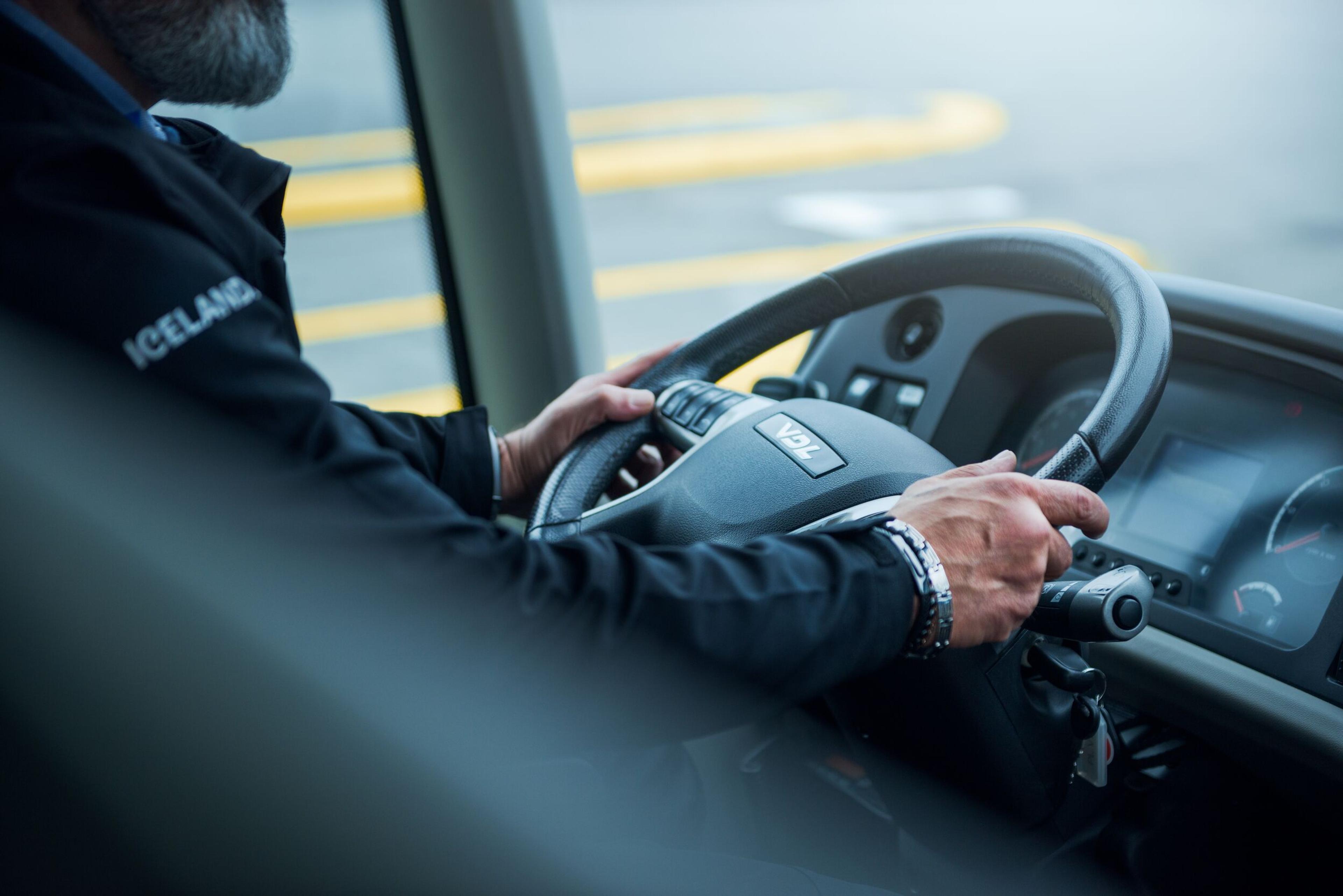 : Close-up of a driver holding the steering wheel of a tour bus in Iceland, wearing a jacket with "Iceland" written on the sleeve.