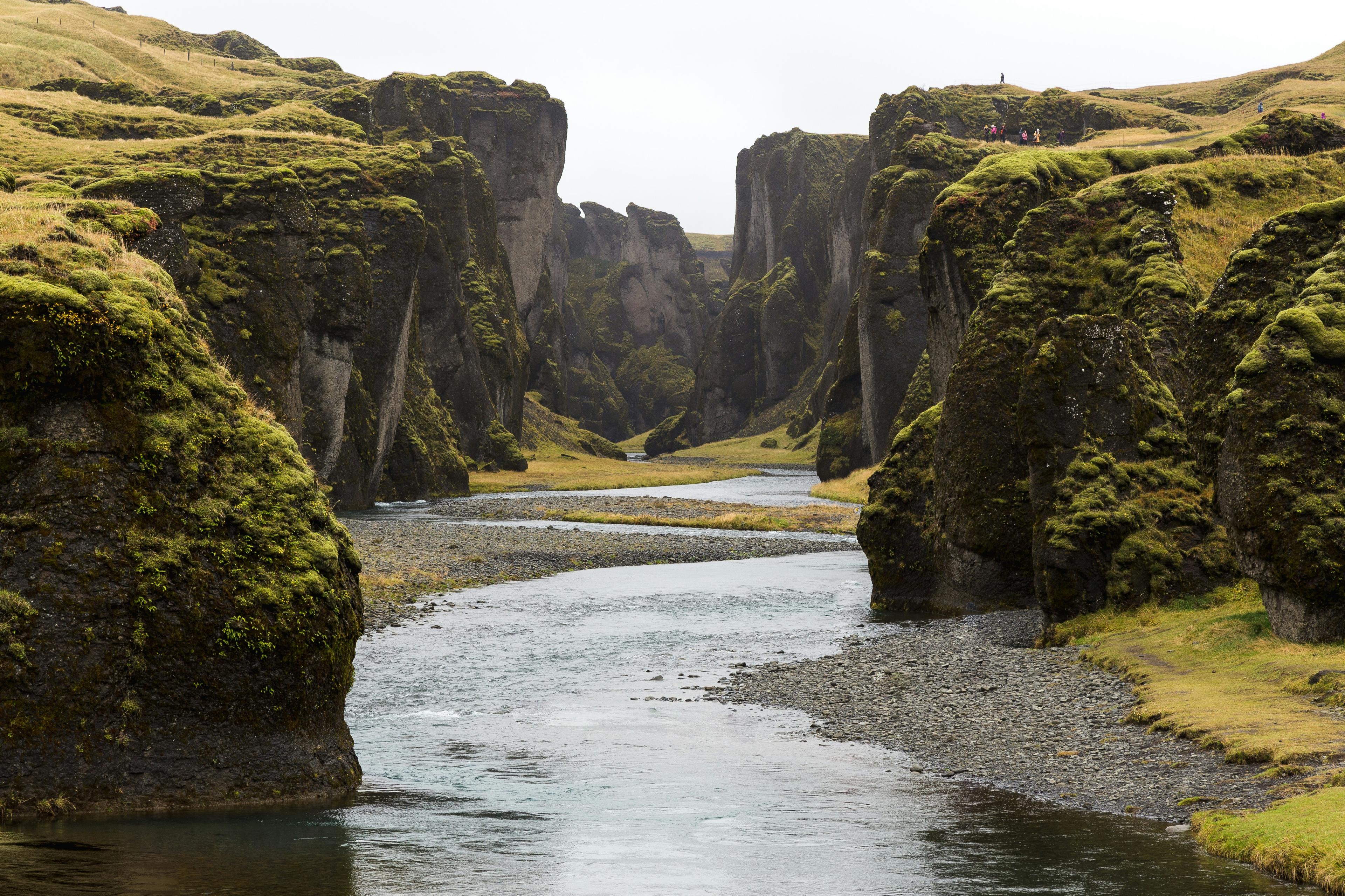 A tranquil river meandering through a dramatic, moss-covered canyon under a cloudy sky.