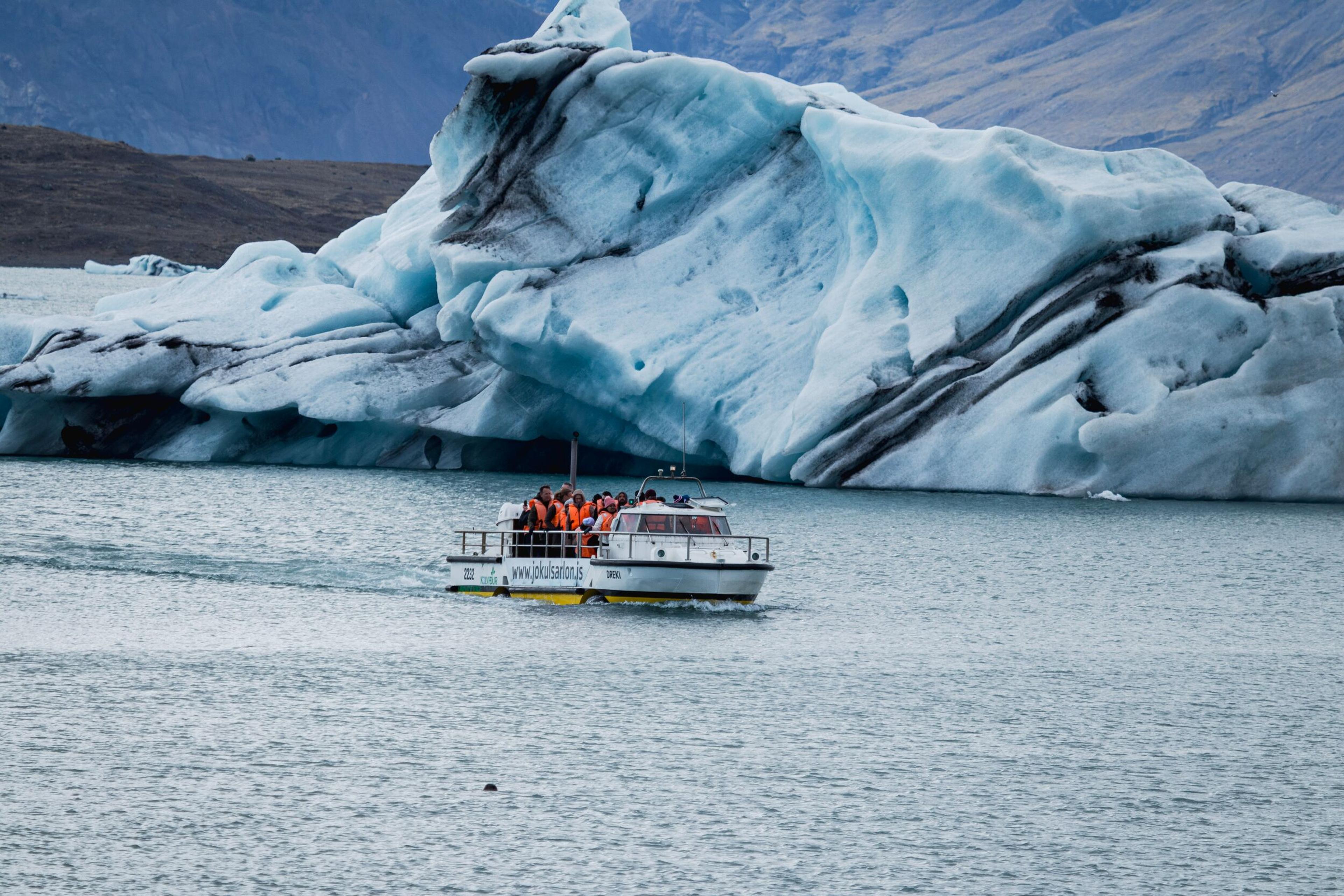 A tour boat filled with people in orange life vests moves through a glacial lagoon past enormous blue and white icebergs.