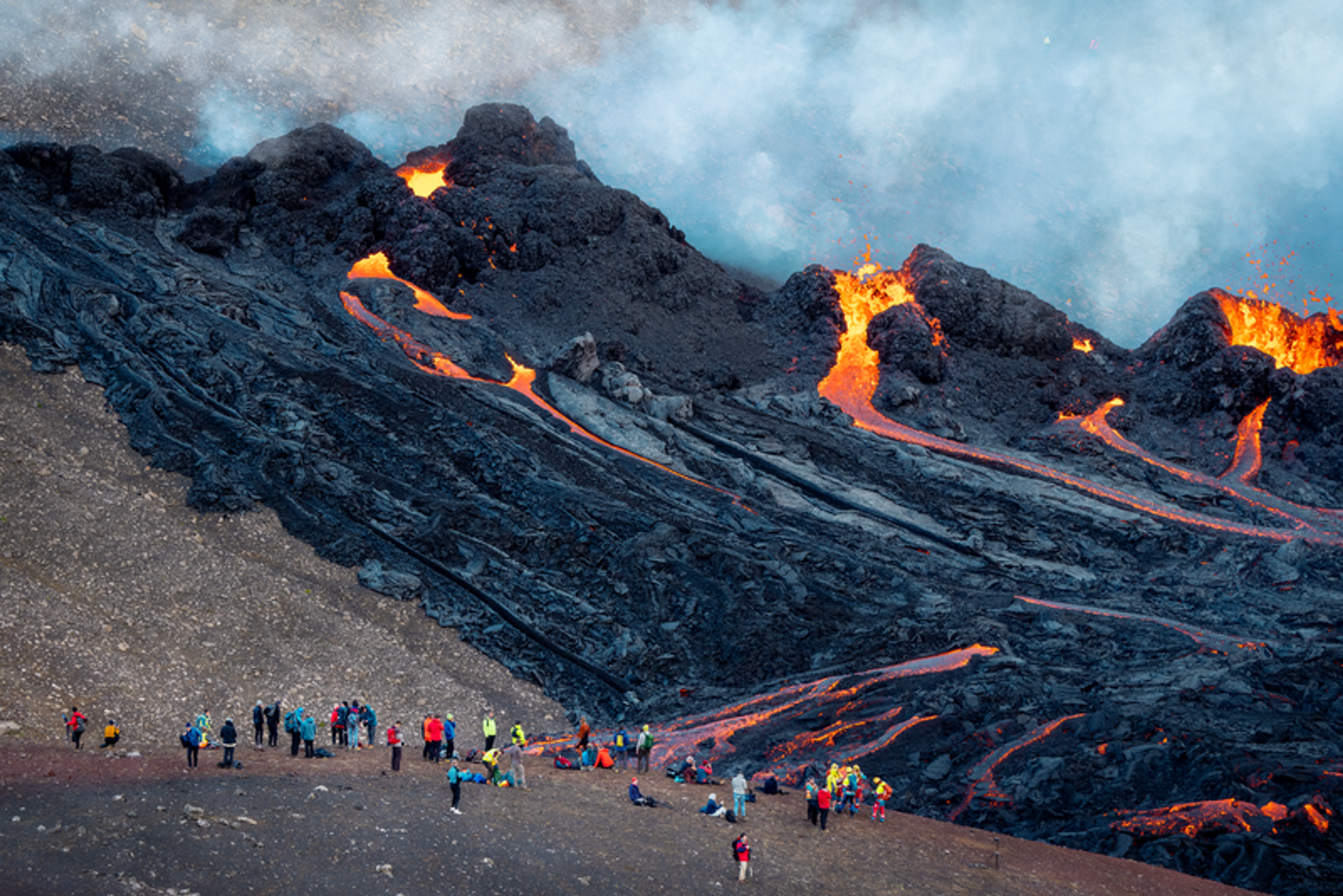 Spectators watching rivers of lava flowing from volcanic fissures on a dark, rugged terrain.