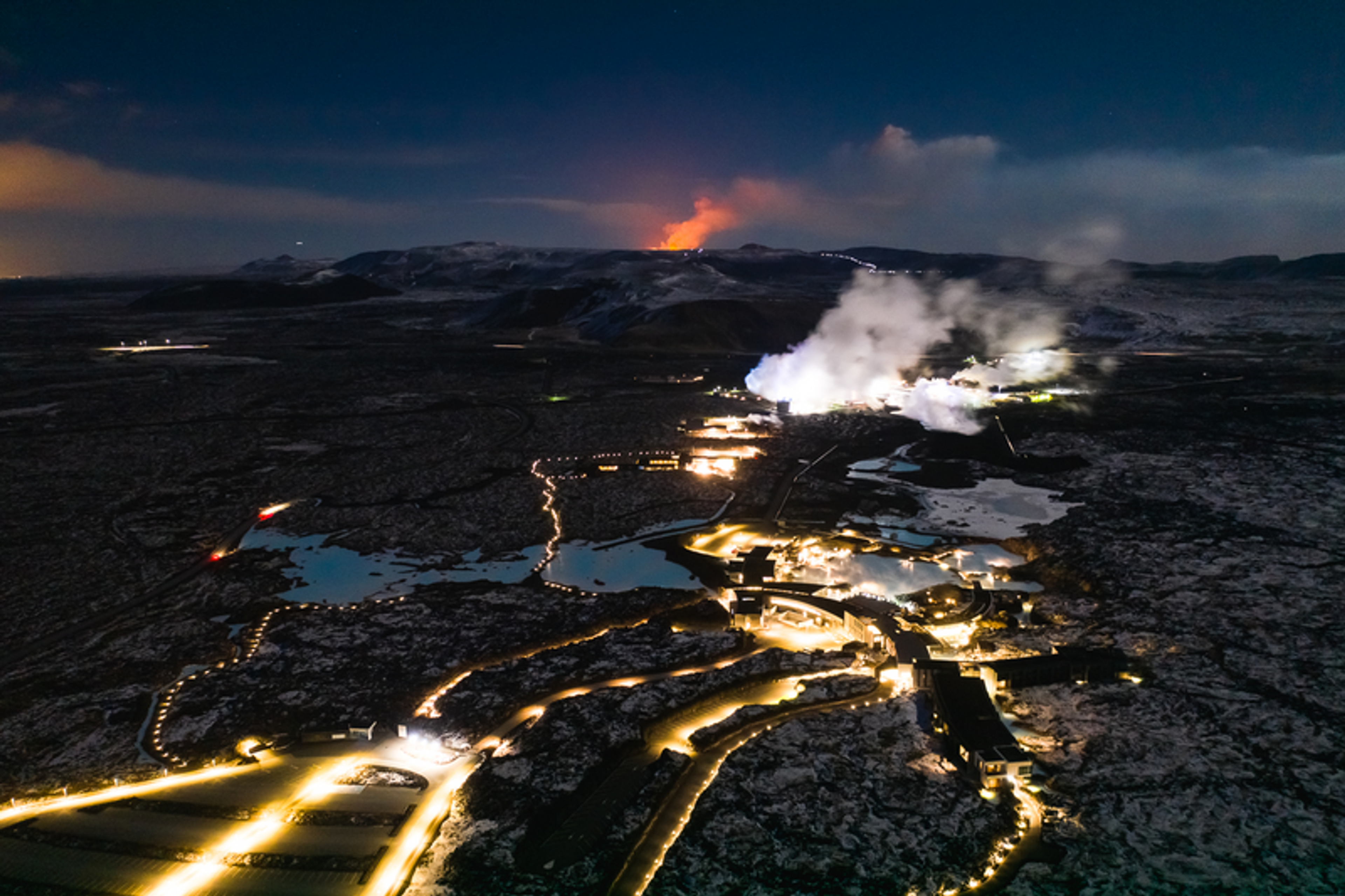 Aerial night view of Fagradalsfjall volcano erupting in the distance, with smoke rising and illuminated geothermal pools and buildings in the foreground.