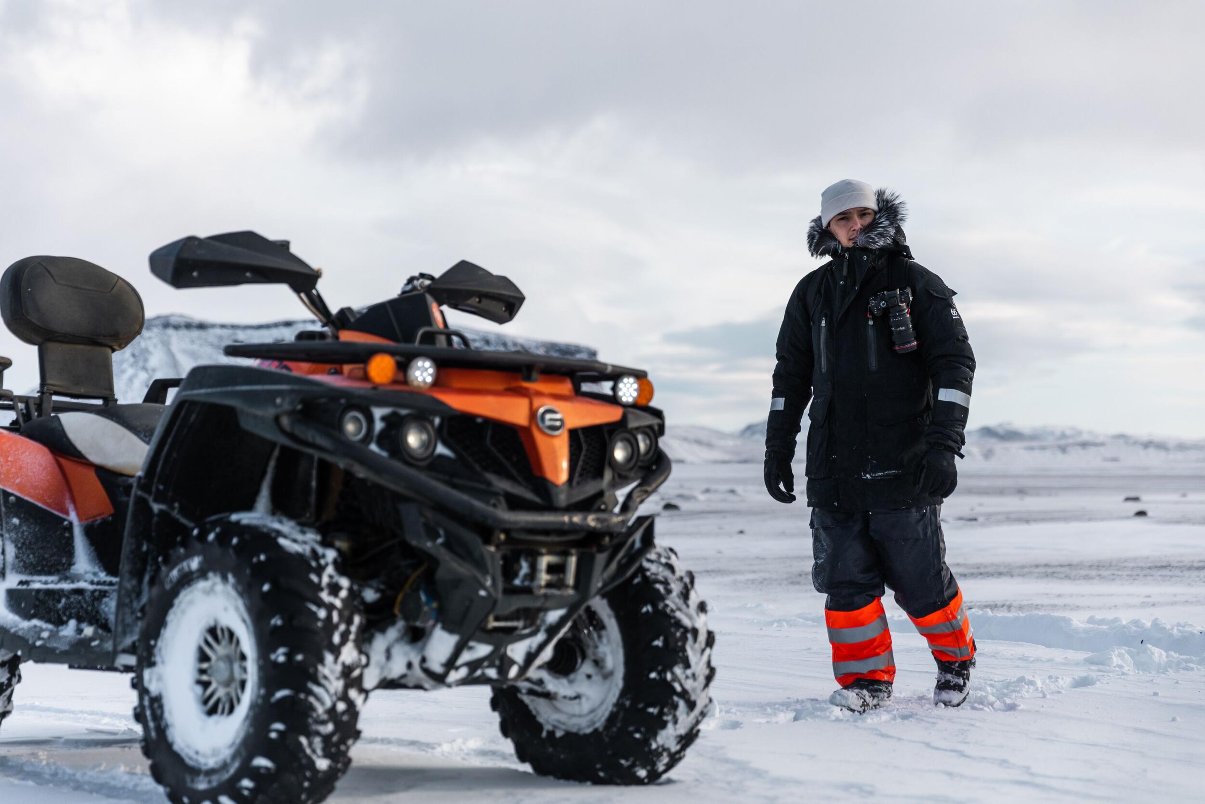 A rugged ATV covered in snow is parked on an icy landscape, with a warmly dressed tour guide in heavy winter gear standing beside it, set against Iceland’s frozen wilderness.