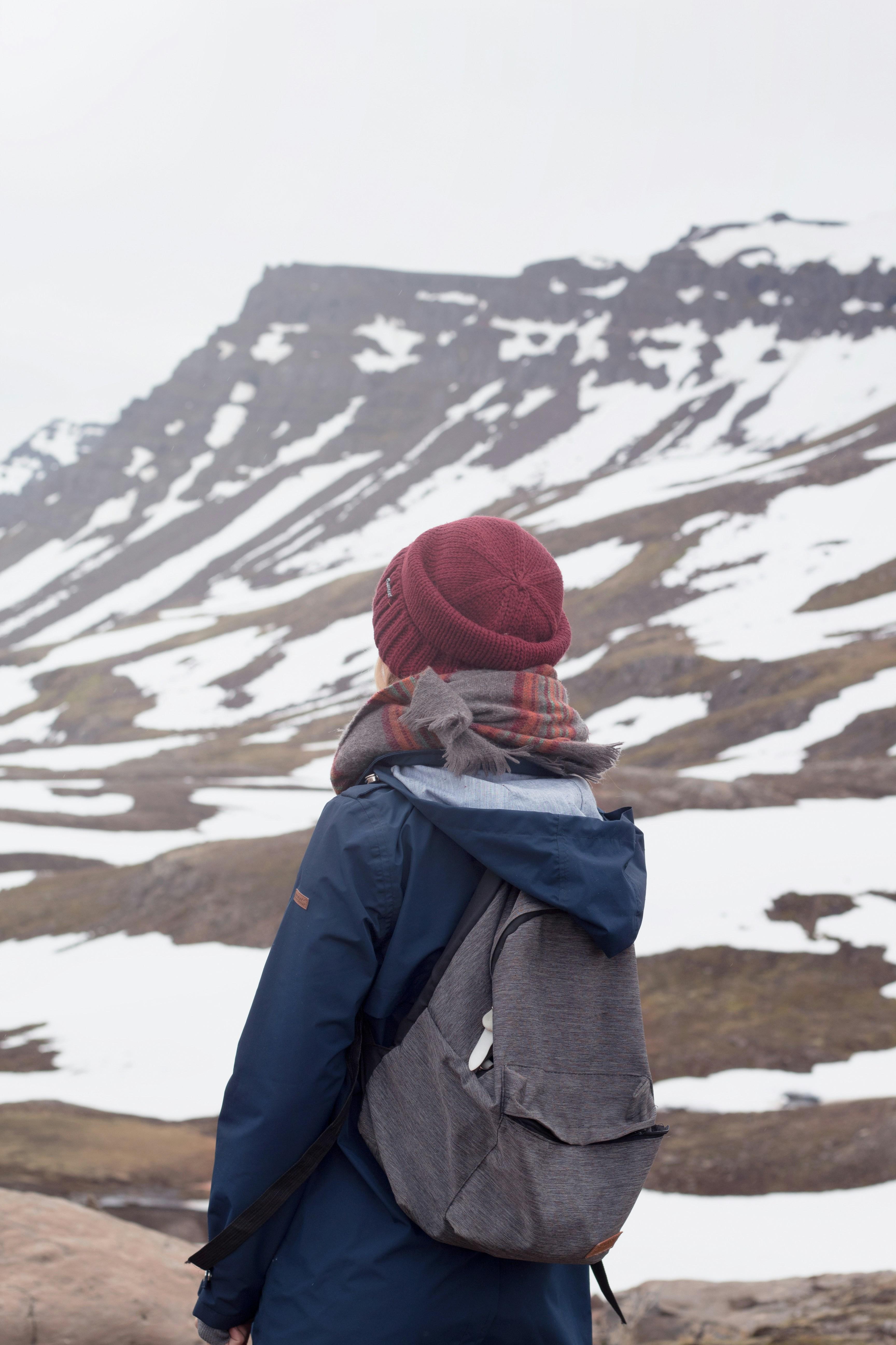 A person wearing a red knit hat, scarf, and backpack gazing at a snowy mountain landscape with patches of bare ground and overcast skies