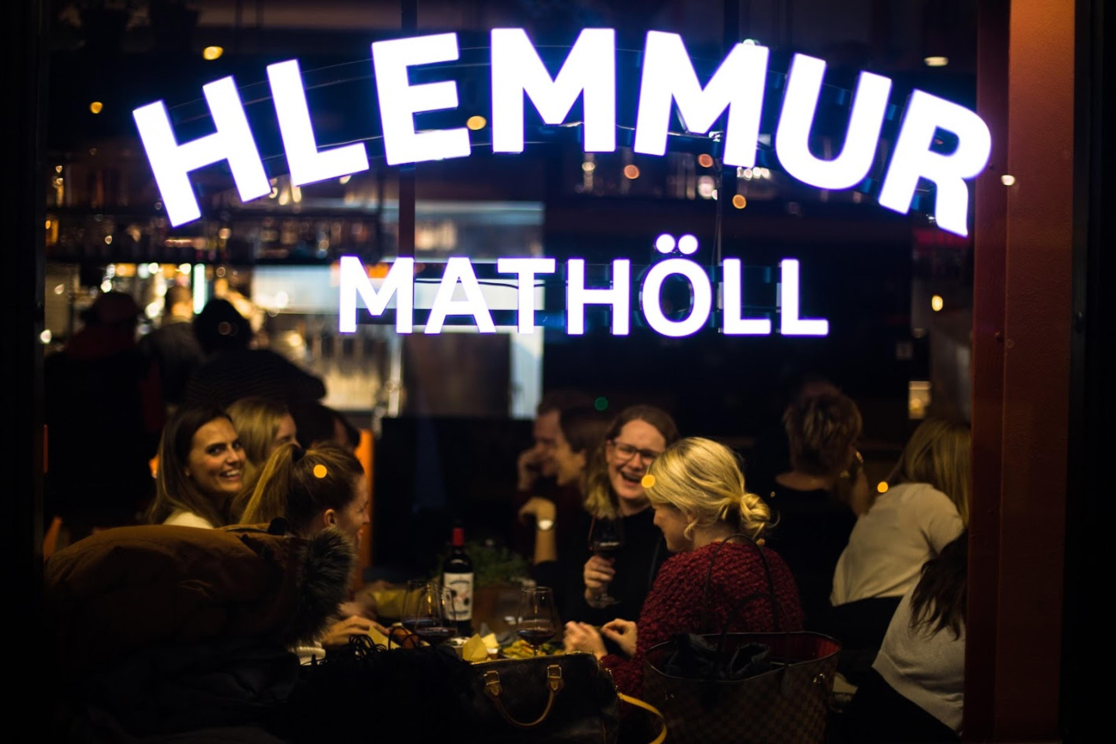 People enjoying a lively evening at Hlemmur Mathöll, seen through the window with the bright sign above, sharing food and drinks in a warm, social atmosphere.