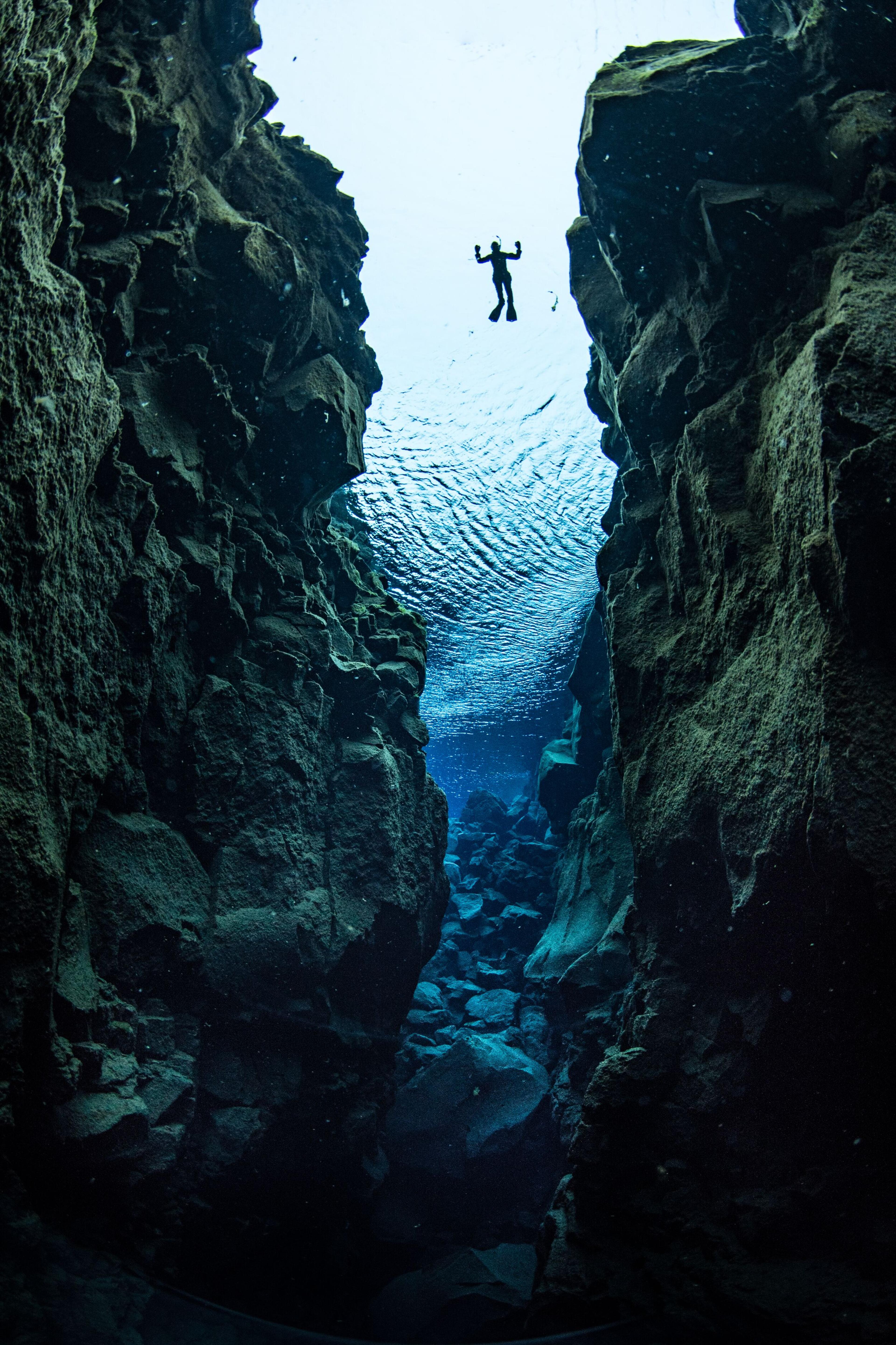 A silhouette of a diver suspended in the clarity of the water, between the narrow walls of a rock fissure, with light penetrating from the surface, highlighting the serene and majestic underwater environment of Silfra.