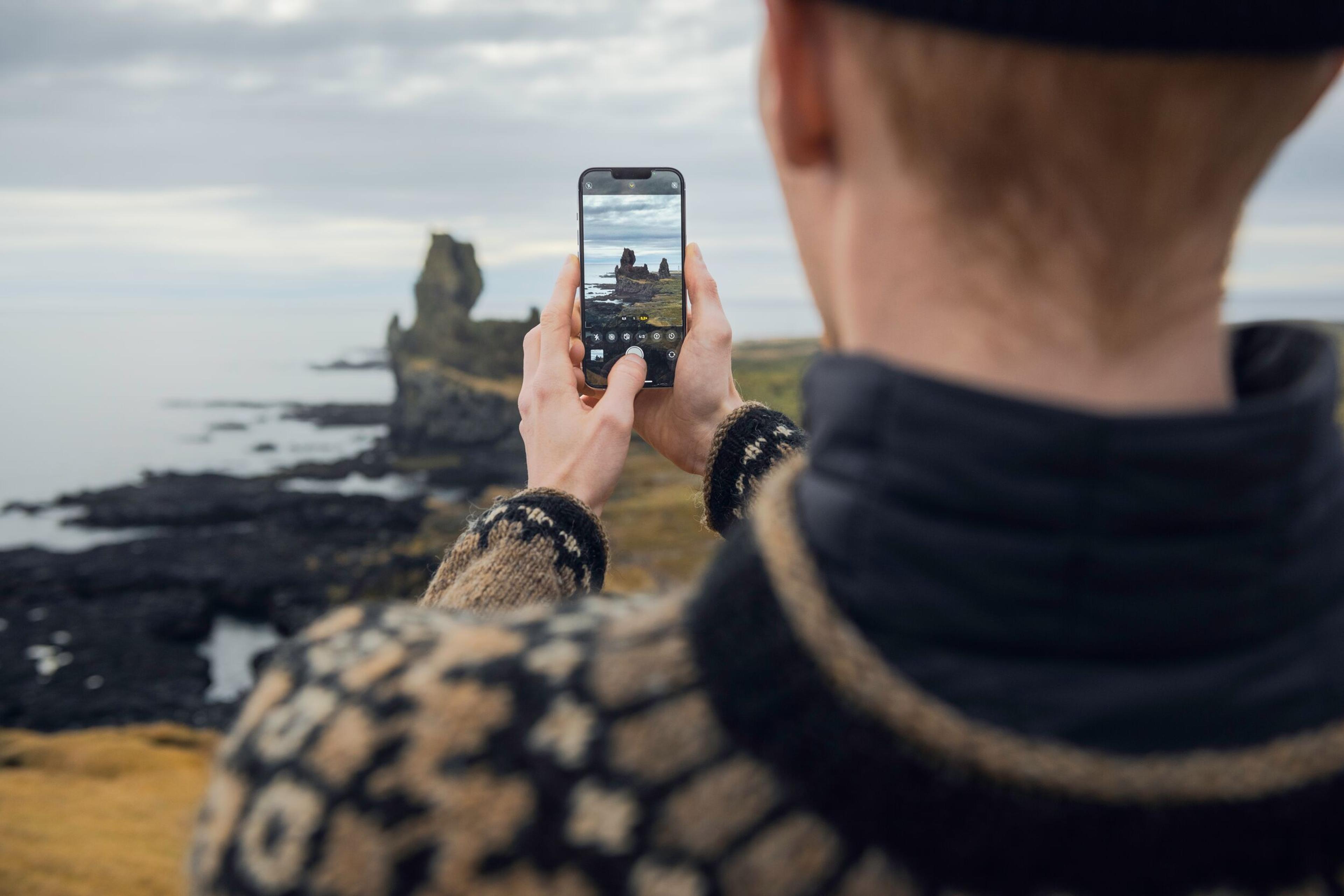 A man using his phone to capture a photo of a prominent sea stack.