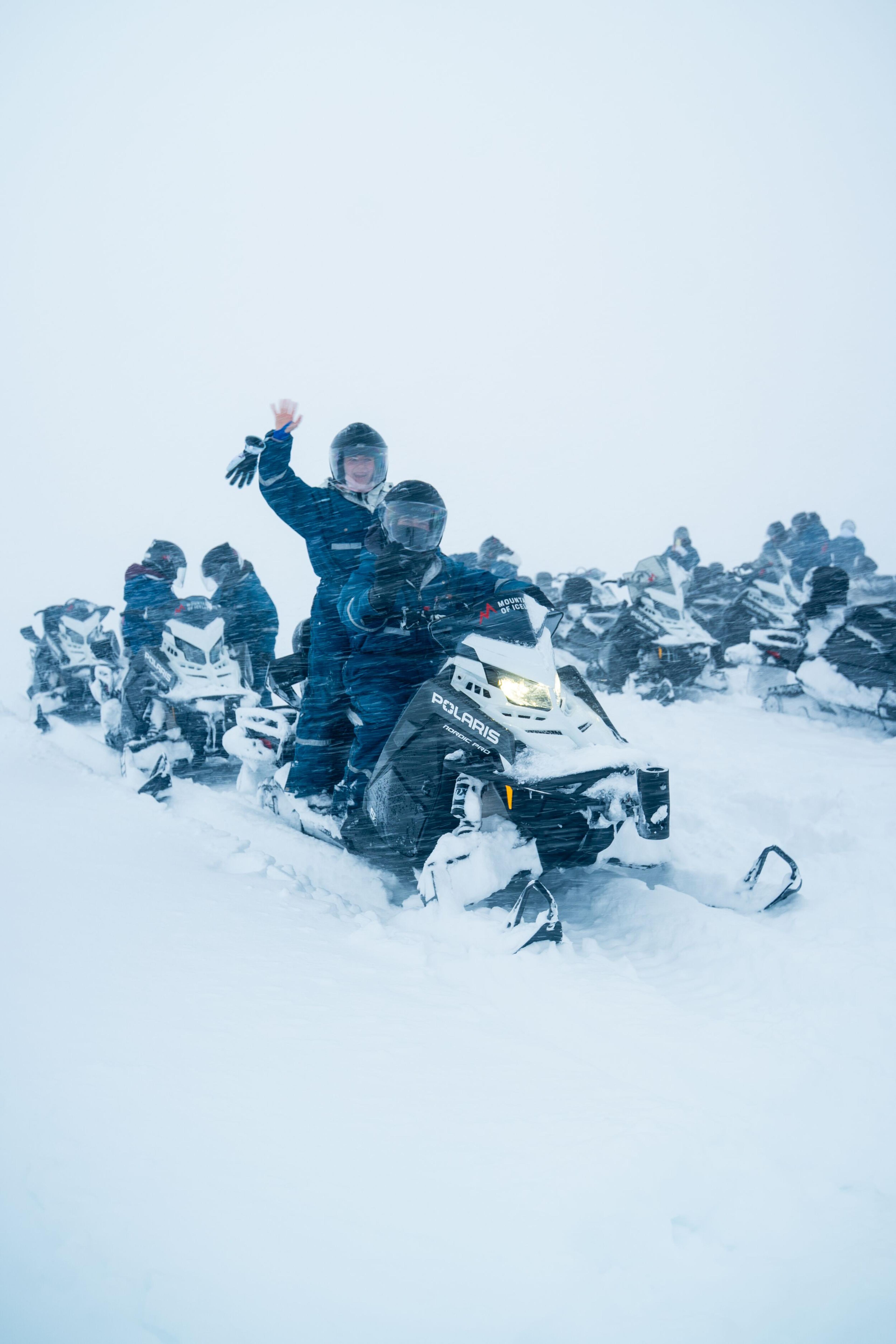 A group of people on snowmobiles in heavy snowfall, with one person waving from the front.