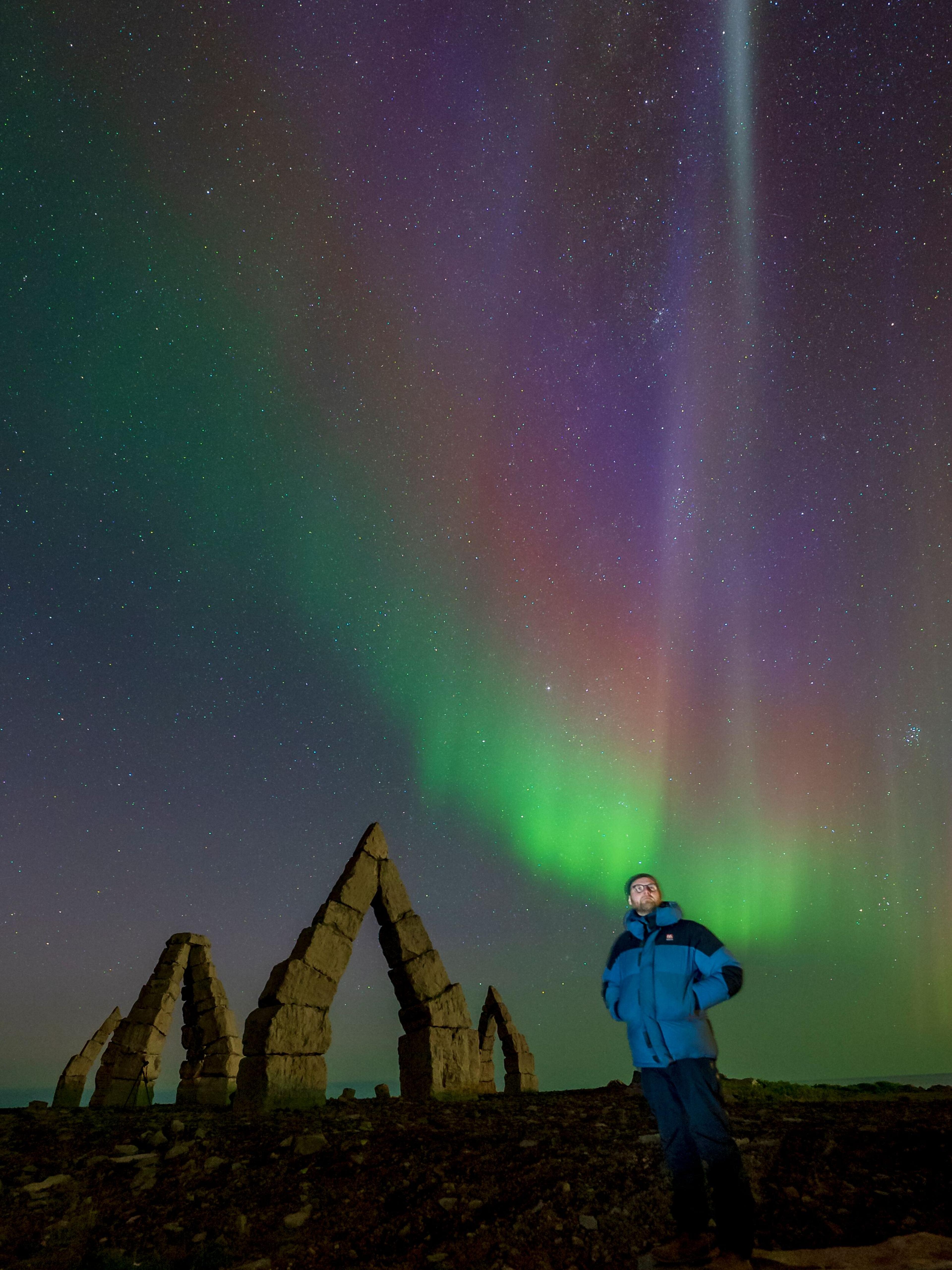 A person in a blue jacket stands near a stone archway, with vibrant green and purple Northern Lights illuminating the night sky.