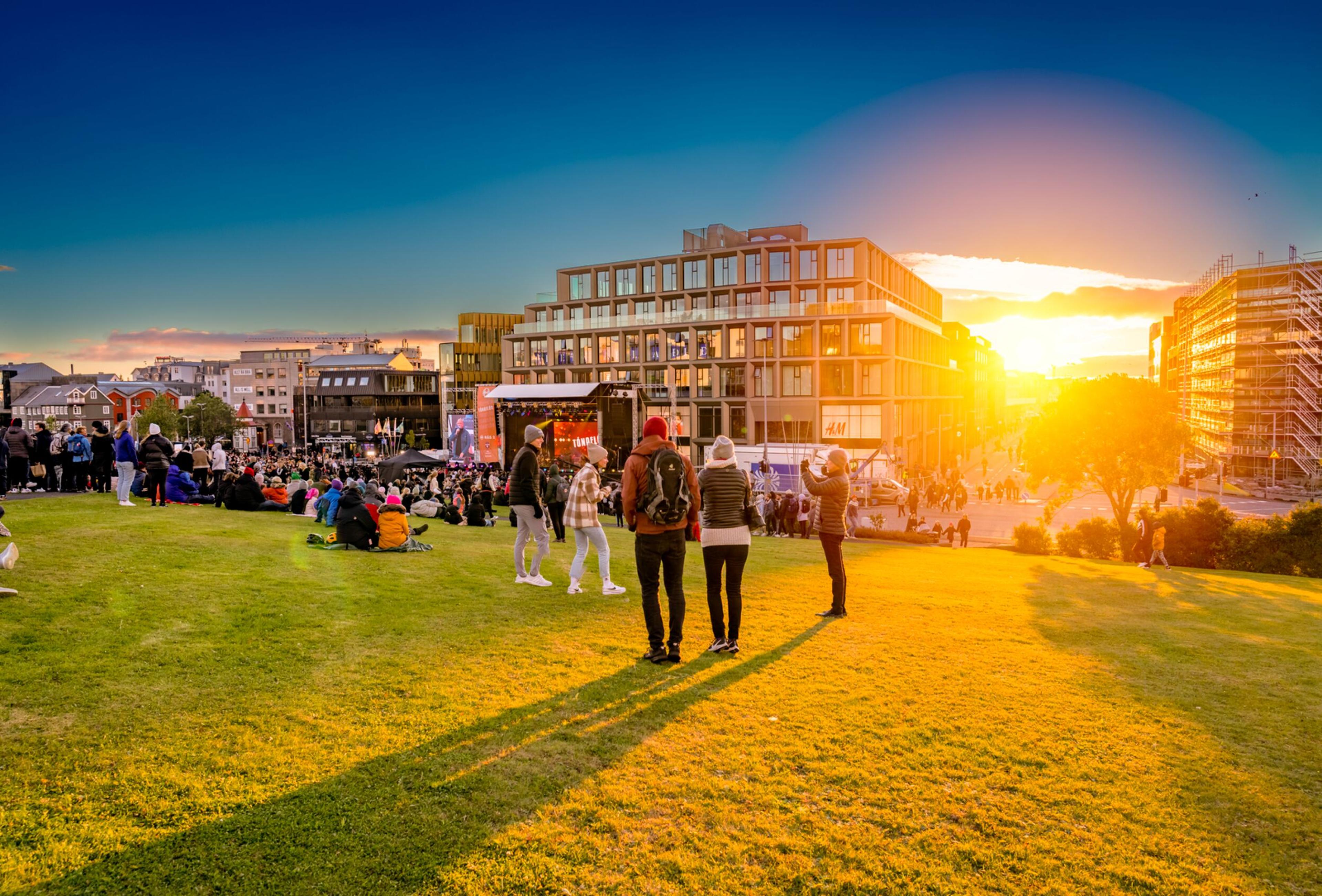 People gather in a sunlit park with modern buildings in the background, enjoying an outdoor event during Reykjavík Culture Night at sunset.