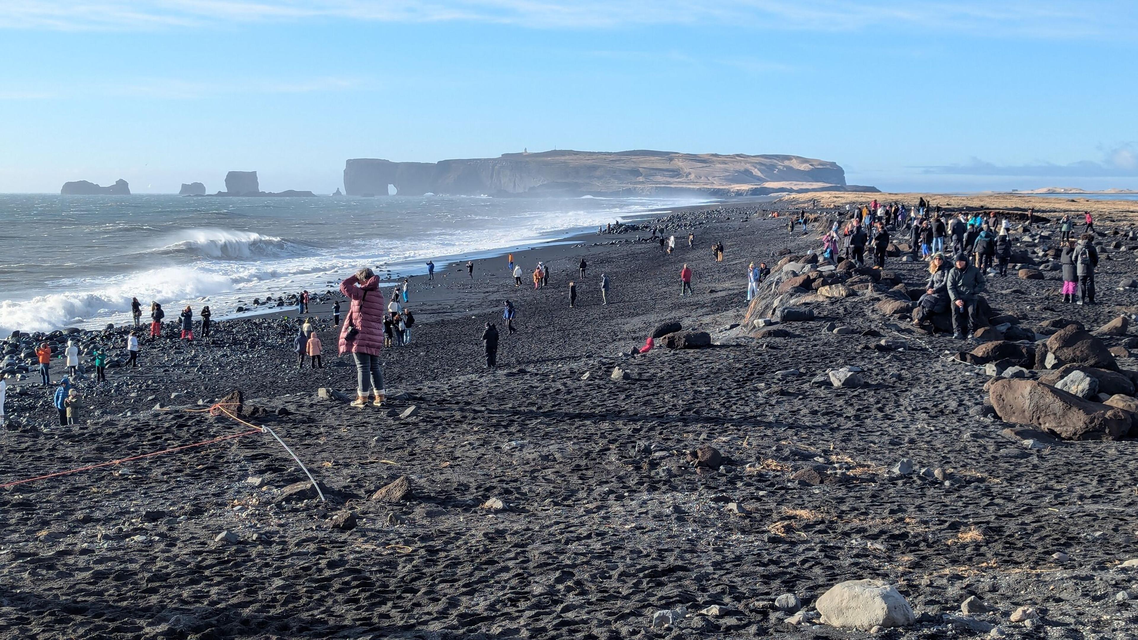 A crowded black sand beach with crashing waves and distant sea stacks and cliffs.