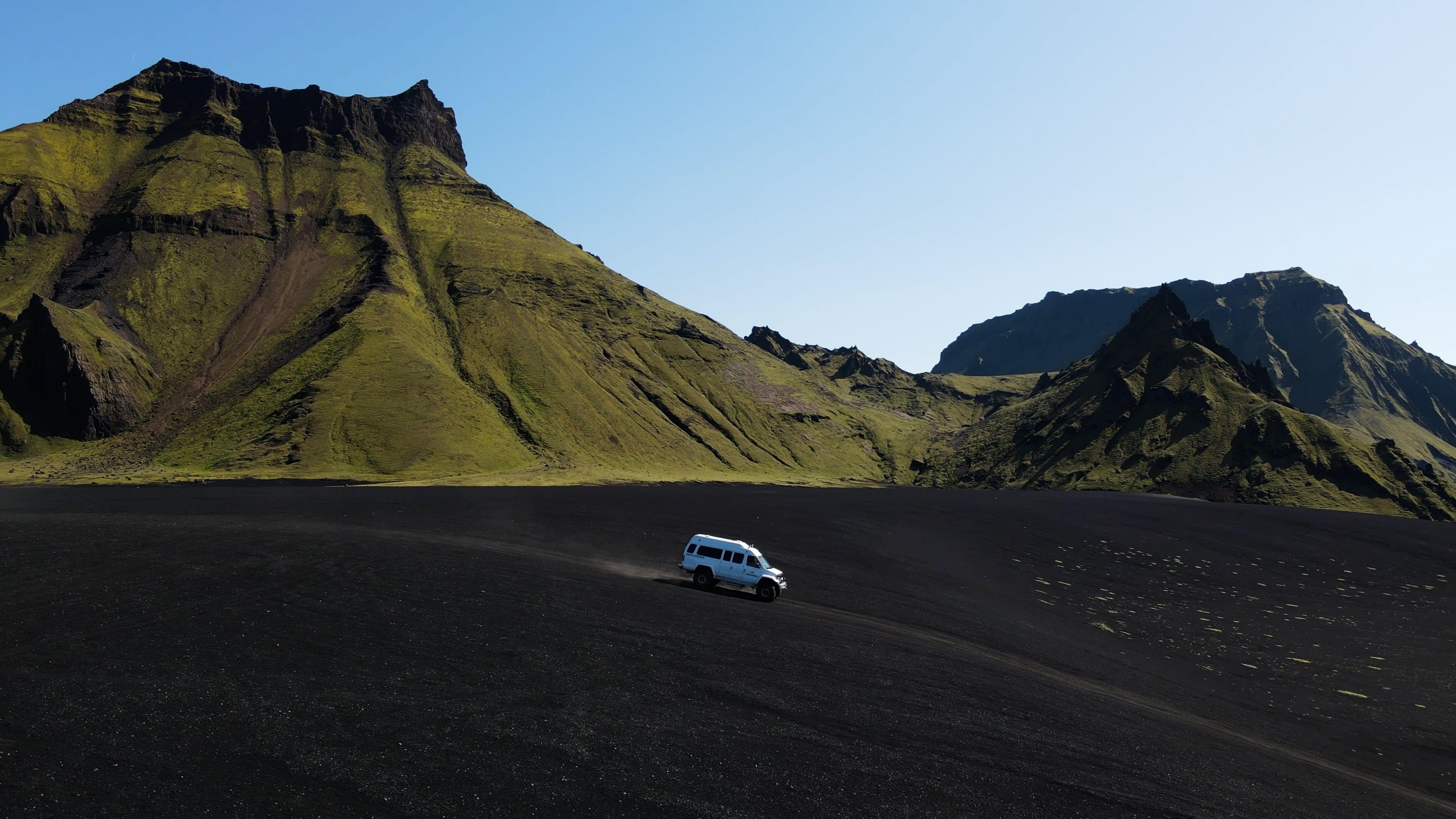 A white off-road vehicle drives across a black volcanic plain with green mountains under a blue sky.