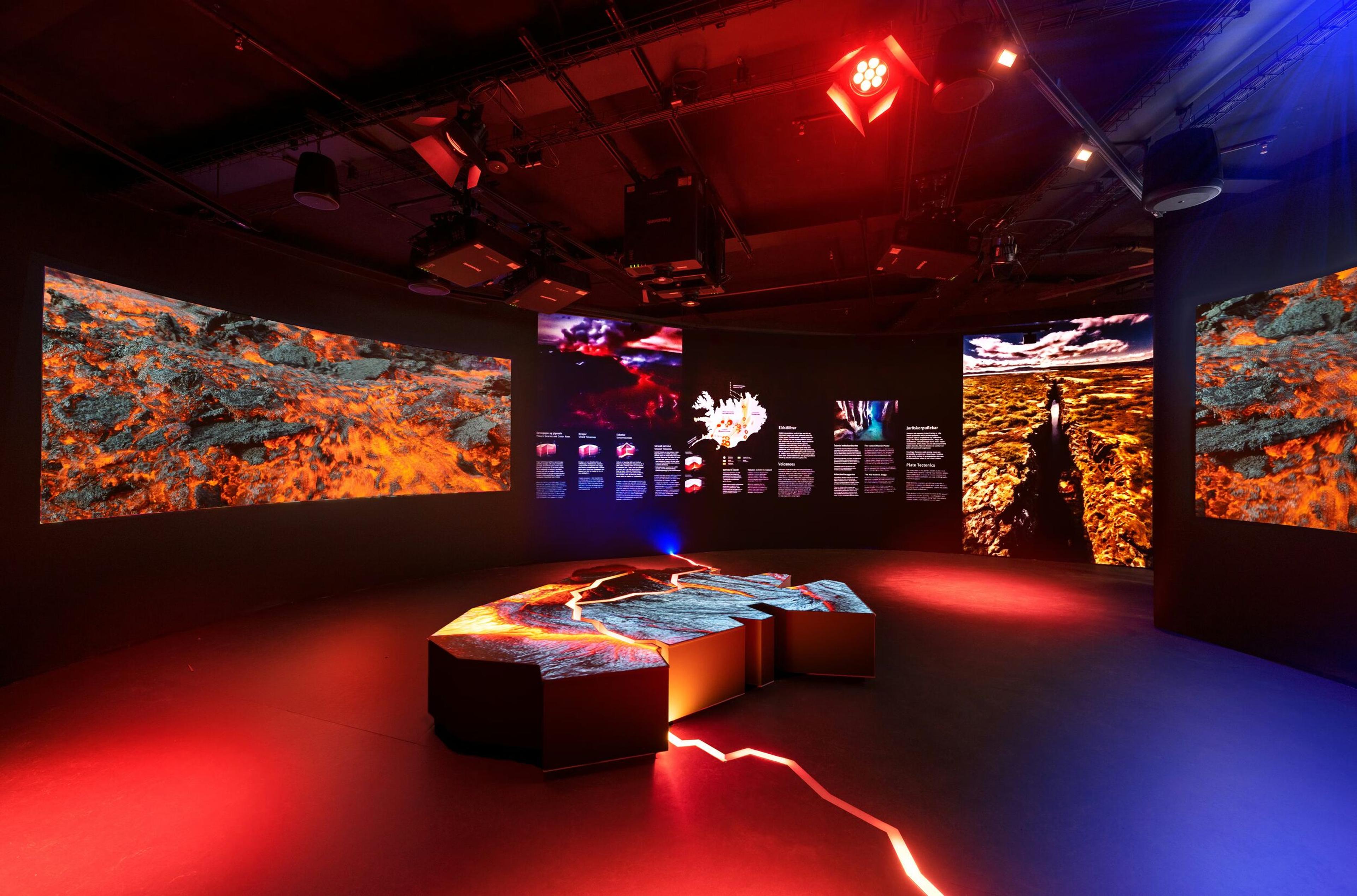 A dark exhibition room with large screens displaying volcanic landscapes and lava, a central illuminated table projecting lava, and red and blue mood lighting.