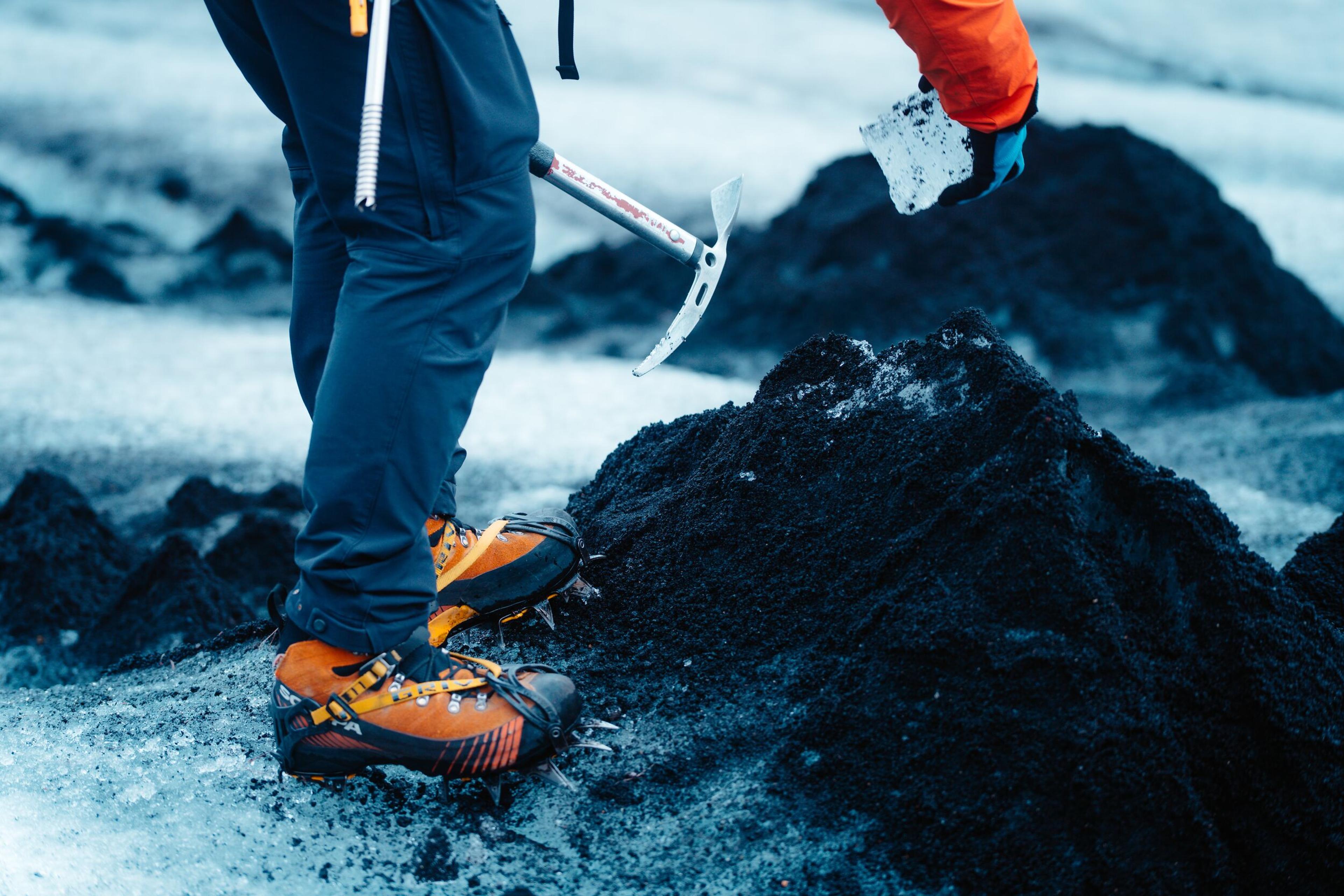 A person wearing mountaineering boots and crampons stands on a glacier covered with volcanic ash, holding an ice axe and a piece of ice.