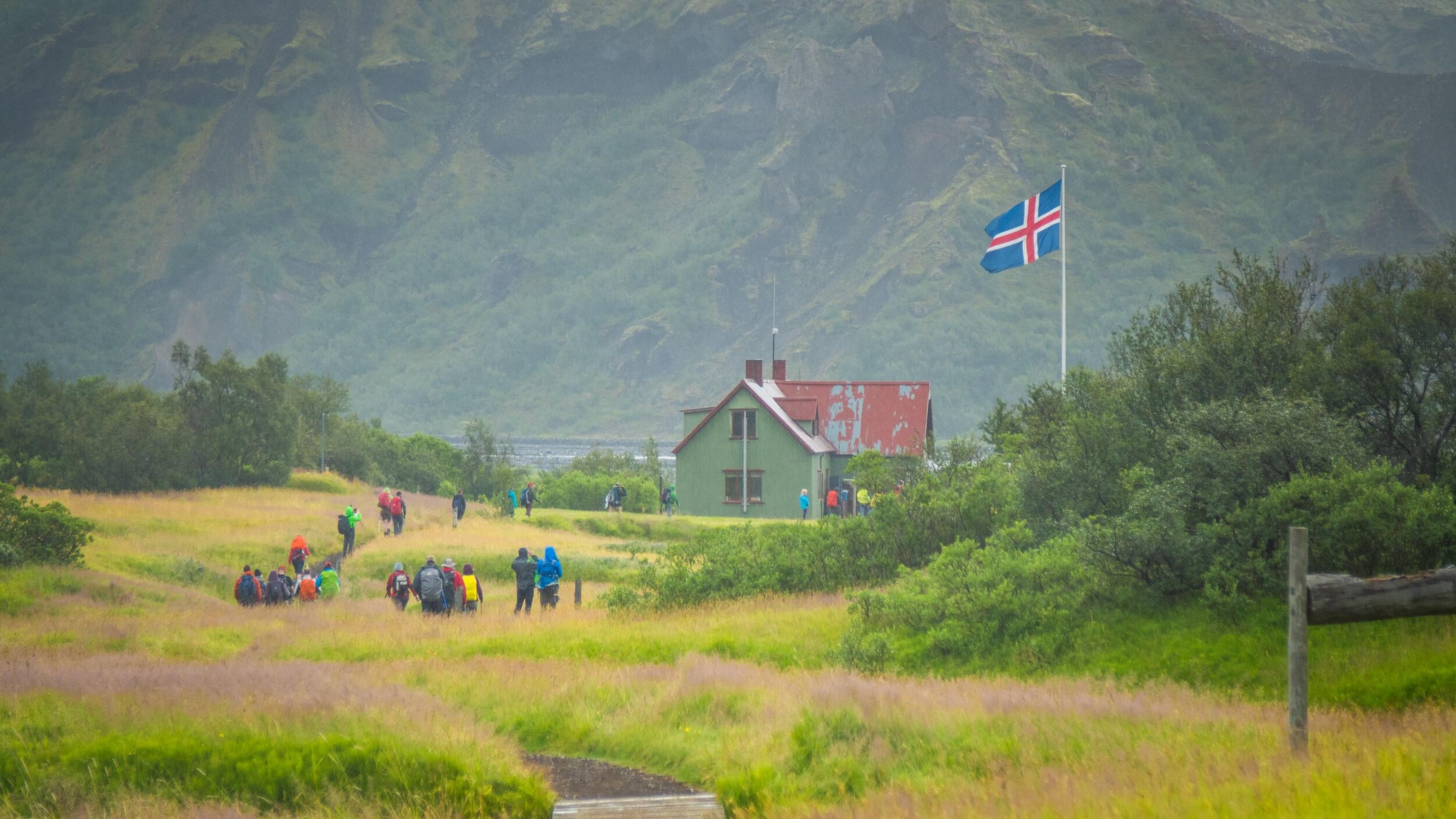 Hikers approaching a mountain hut, distinguished by an Icelandic flag, nestled in a verdant valley.