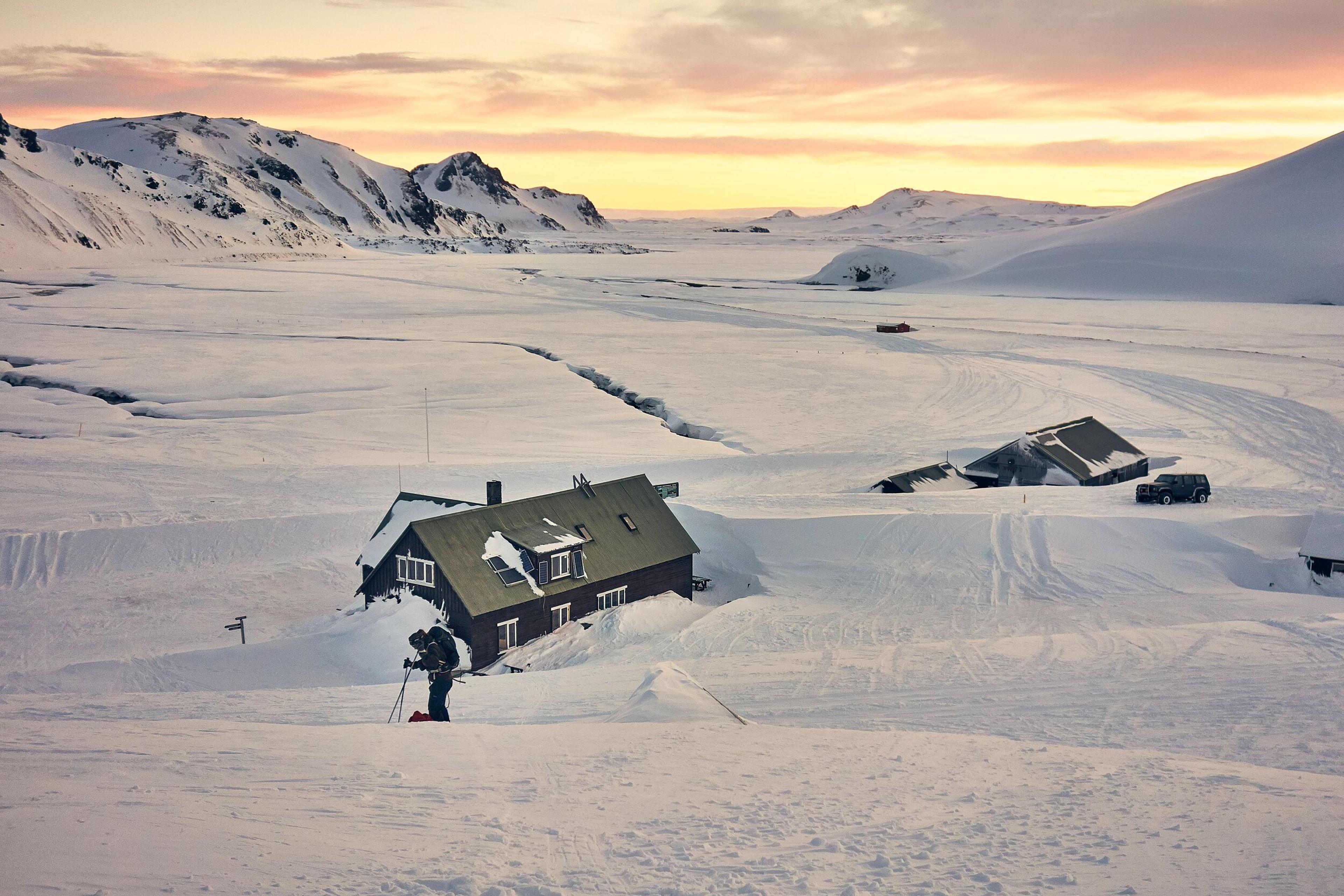 A person on skis stands in deep snow next to a partially buried lodge in a vast, snow-covered mountain landscape under a warm sunset sky.