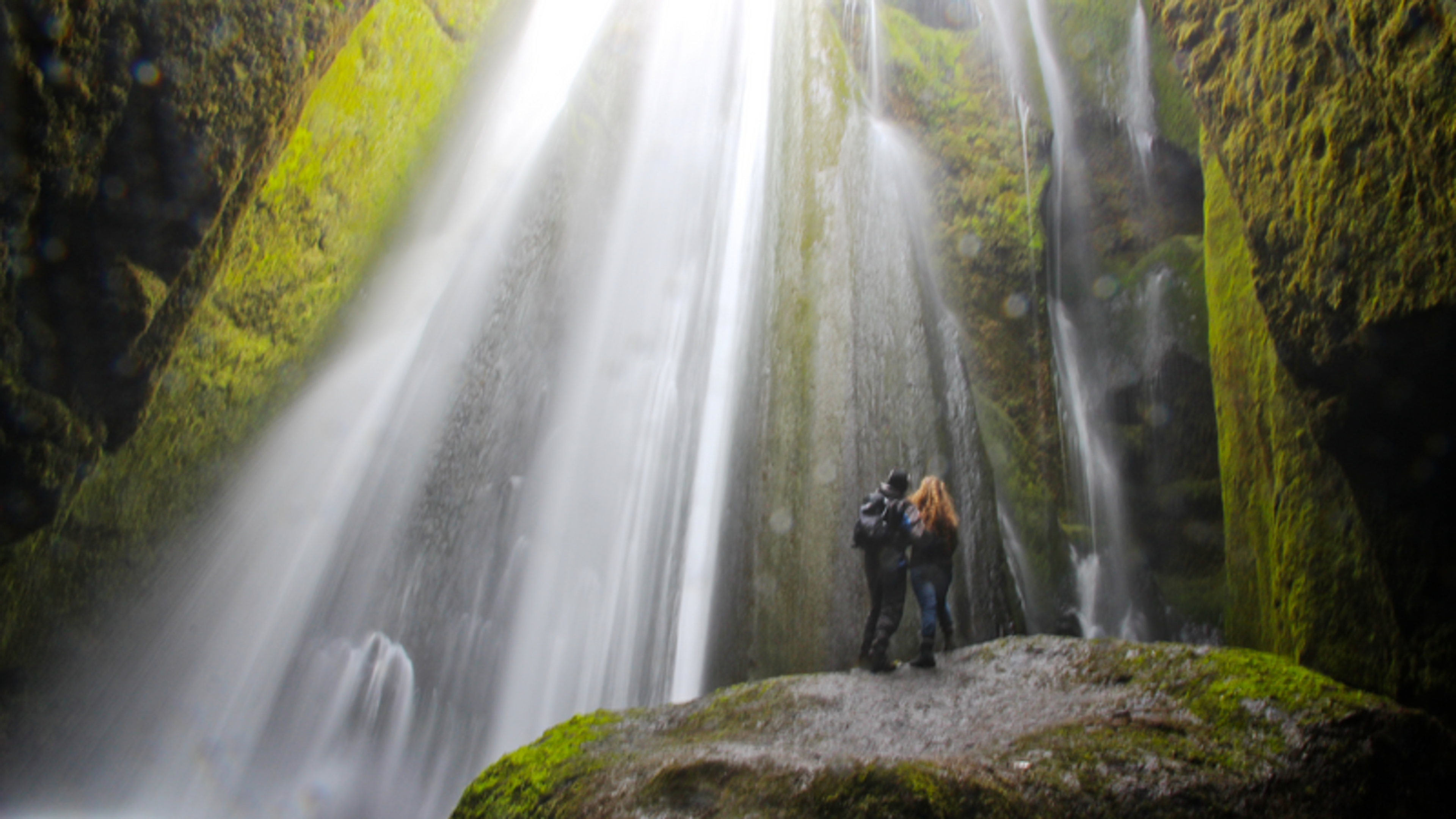 Two people standing on the top of a large rock in a mossy gorge, looking up to the waterfall that's cascading from above and surround them