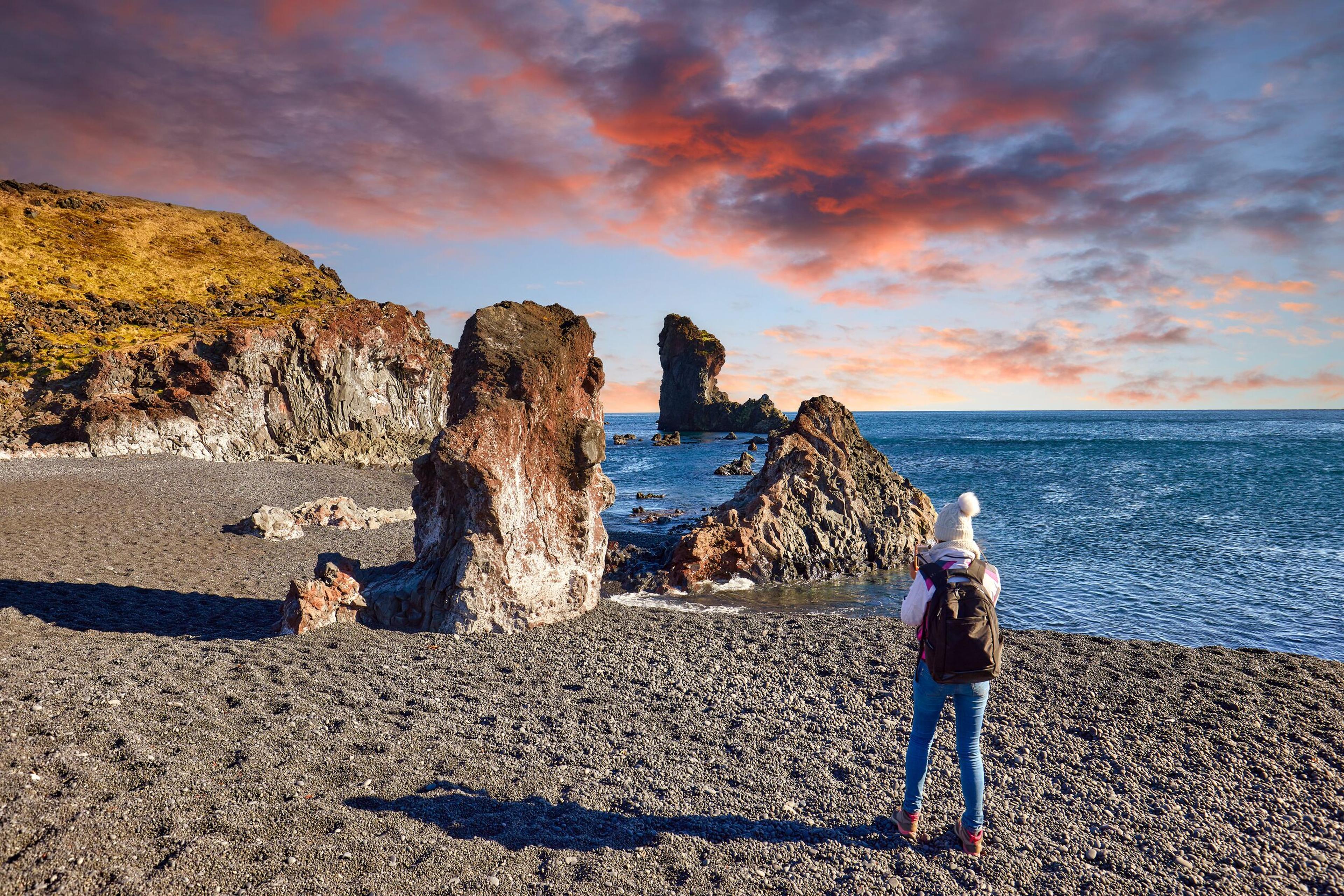 A person observes the dramatic volcanic rock formations along a black pebble beach under a fiery sunset sky, highlighting the rugged coastal beauty.